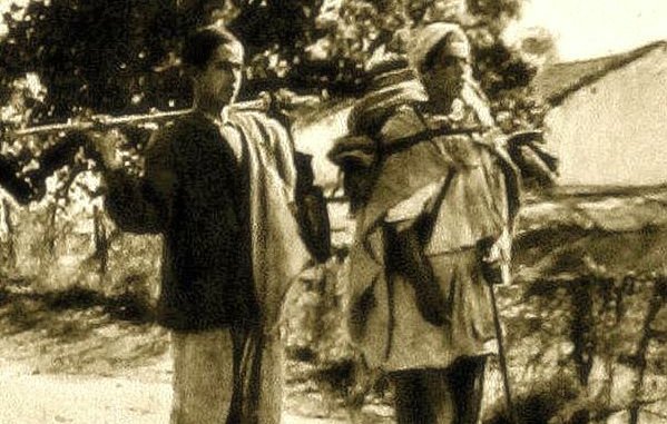 L'image montre deux personnes marchant sur un chemin. L'une d'elles est un jeune garçon, tandis que l'autre est une femme plus âgée. Les deux portent des charges sur leurs épaules, indiquant qu'ils transportent des provisions ou des biens. Le décor semble rural, avec quelques arbres en arrière-plan, et l'atmosphère générale donne un aperçu de la vie quotidienne. Le style de vêtements et le traitement de la photo évoquent une époque passée.