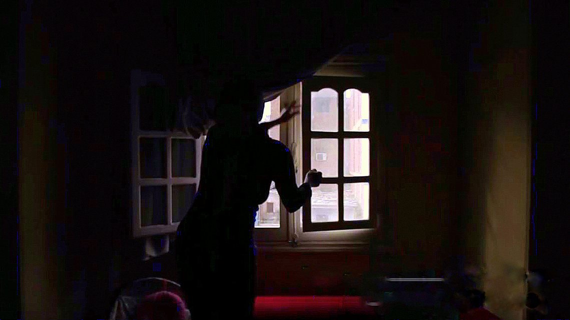 L'image montre une silhouette se tenant près d'une fenêtre dans une pièce sombre. La lumière entre par la fenêtre, illuminant partiellement l'espace environnant, tandis que des détails tels que les murs et les meubles sont à peine visibles dans l'ombre. La silhouette semble en mouvement, ajoutant une ambiance dynamique et mystérieuse à la scène. Les rideaux sont légèrement ouverts, laissant entrevoir l'extérieur.