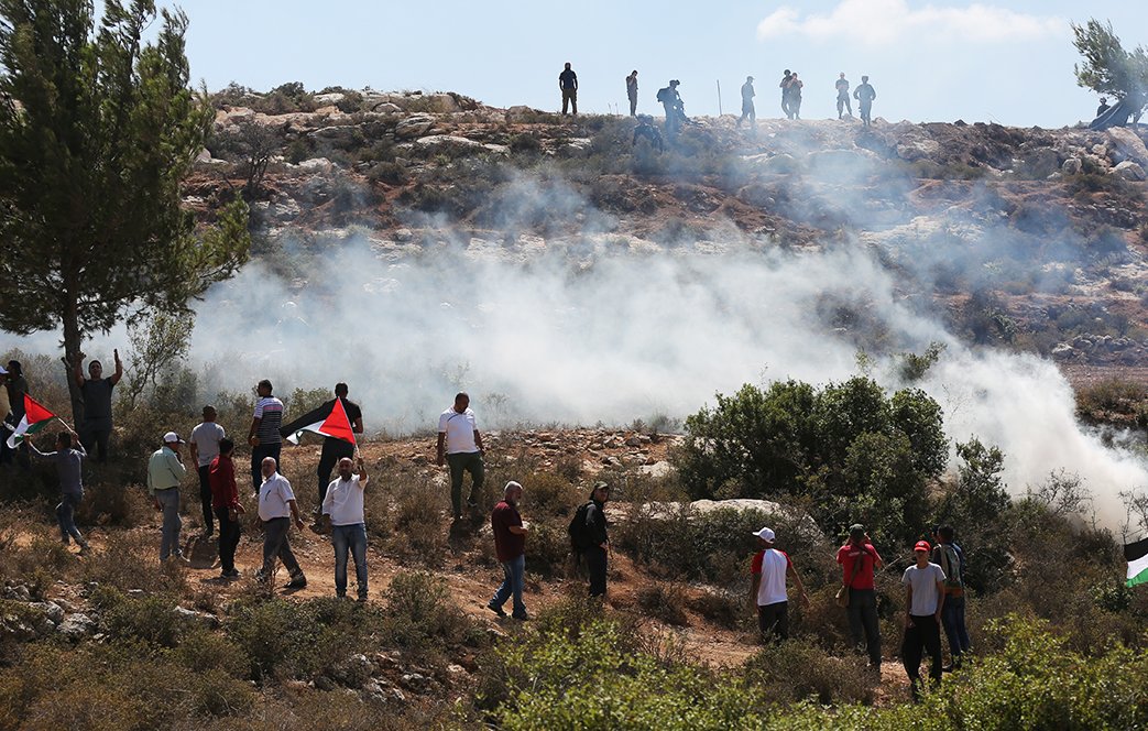 The image depicts a scene with a group of people gathered in an outdoor setting, likely demonstrating or protesting. In the foreground, some individuals are waving flags, possibly representing Palestine, characterized by red, black, and green colors. Behind them, a cloud of smoke rises, suggesting the use of tear gas or other crowd control measures. In the background, a group of people can be seen on higher ground, observing the situation. The environment appears to be rocky and natural, with trees scattered in the landscape under a partly cloudy sky.