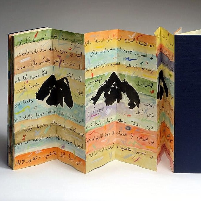 L'image montre un livre artistique aux pages colorées, présentant des illustrations de montagnes. Les pages sont pliées en accordéon et comportent des inscriptions en arabe. La palette de couleurs est variée, avec des nuances de jaune, bleu et orange, ajoutant une dimension vibrante à l'ensemble. Le livre est partiellement ouvert, révélant sa structure unique et sa conception créative. Une couverture noire est visible à droite, contrastant avec les couleurs vives des pages intérieures.