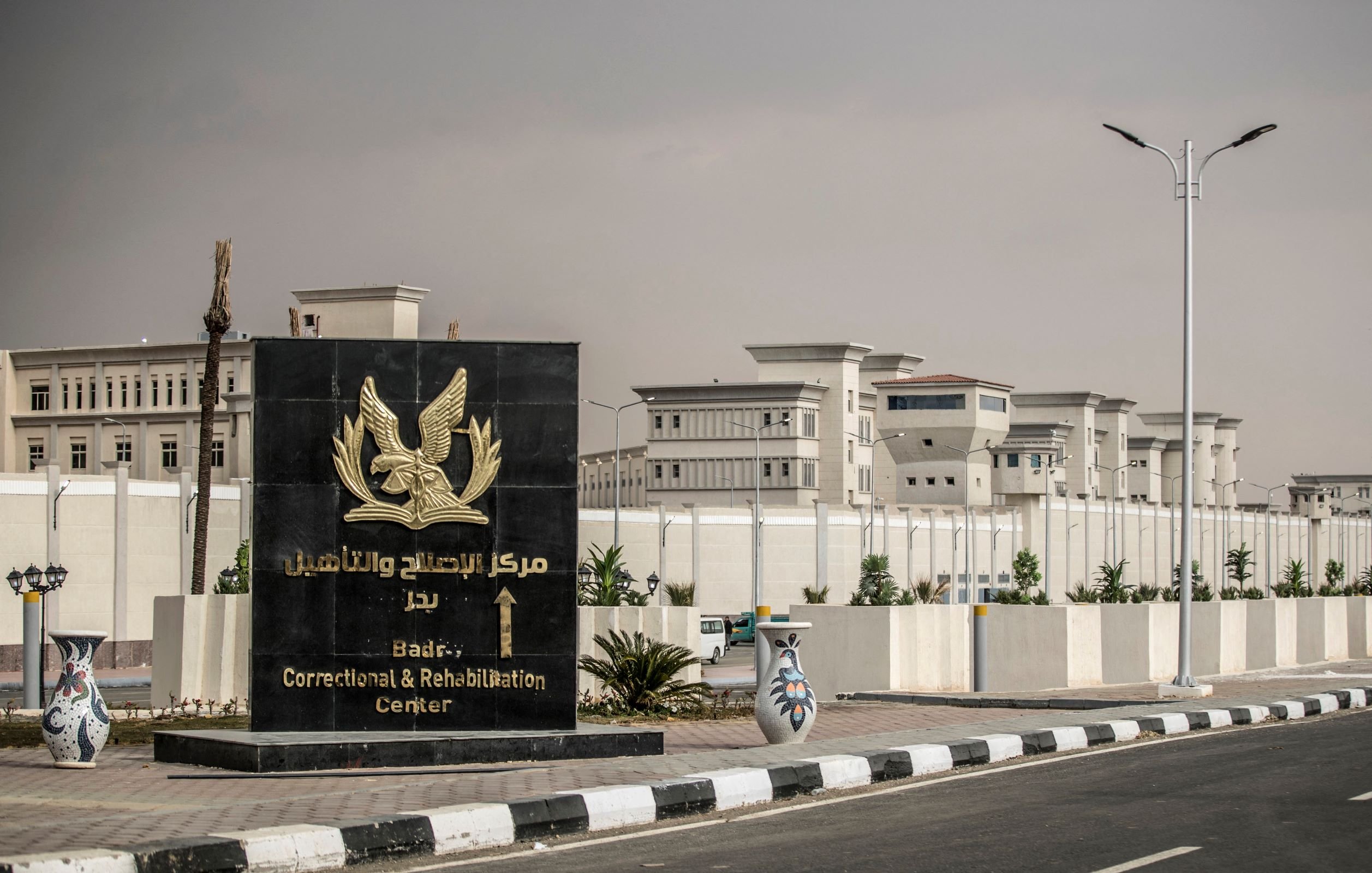 L'immagine mostra un grande cartello di benvenuto che indica il "Centro di Correzione e Riabilitazione di Badia". Il cartello è di colore scuro con un simbolo o un emblema, e si trova all'ingresso di una struttura recintata. Sullo sfondo si possono vedere edifici moderni, circondati da piante e un'illuminazione stradale. Il contesto appare ordinato e ben tenuto, suggerendo un'area istituzionale.