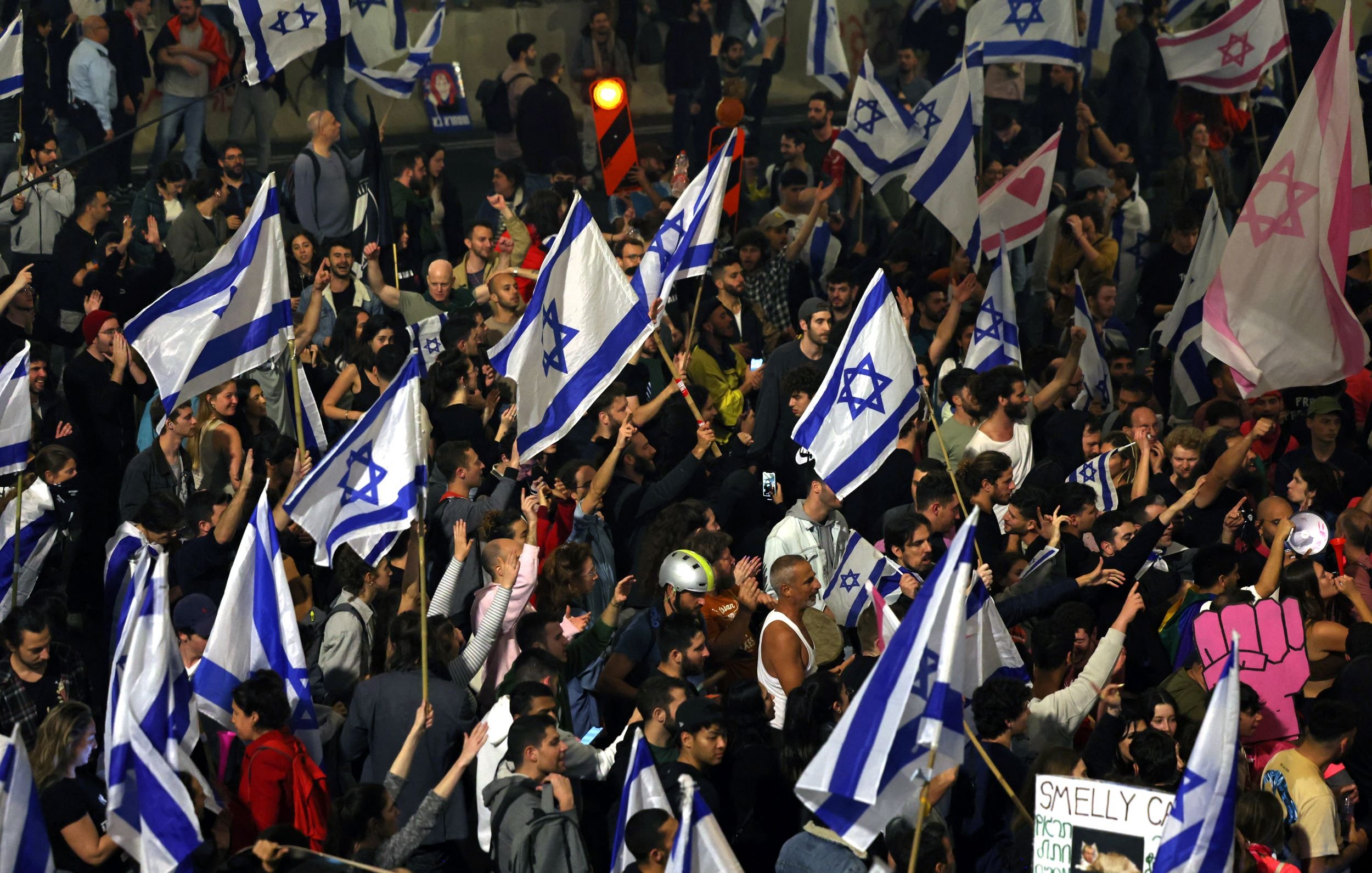 در این تصویر، گروه زیادی از مردم در حال تظاهرات هستند. آنها پرچم‌های آبی و سفید با ستاره داوود را در دست دارند. در میان مردم، برخی تابلوهای نوشته‌دار و علامت‌ها نیز دیده می‌شود. فضای کلی نشان‌دهنده‌ یک تجمع پرشور و فعال است. برخی از شرکت‌کنندگان لباس‌های رنگی و متنوعی پوشیده‌اند و به نظر می‌رسد به یک موضوع خاص اعتراض می‌کنند.