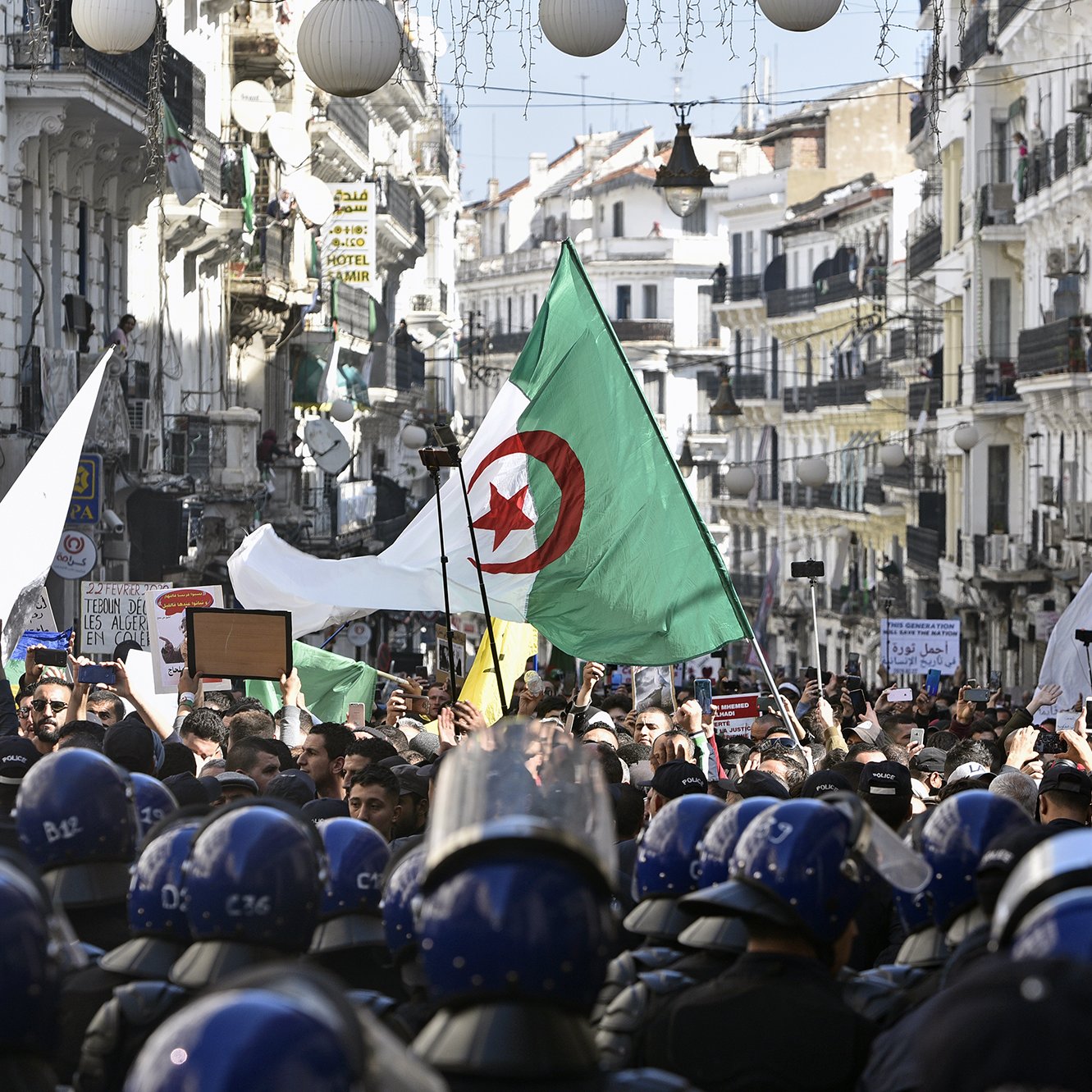 The image depicts a large protest scene in a street lined with buildings. Many people are gathered, holding flags, including Algerian flags, and signs. The crowd is diverse and appears to be demonstrating for a cause. In the foreground, a line of riot police in helmets is visible, standing in front of the protesters, indicating a tense atmosphere. The street is adorned with lights and decorations, suggesting it might be a significant location in the city. Overall, the image captures a moment of civic engagement and protest.