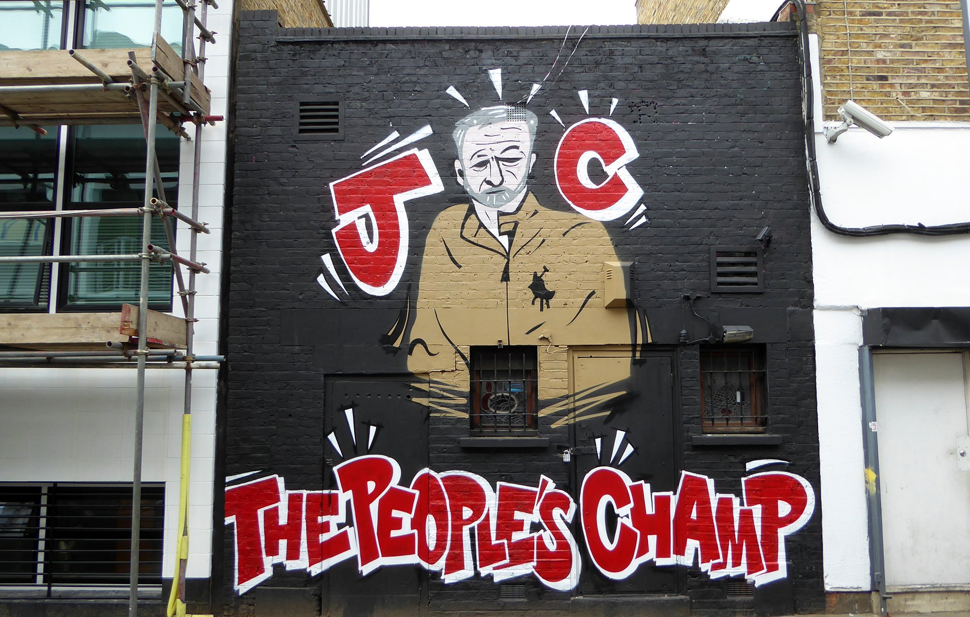 L'image montre un mur décoré d'un graffiti représentant une silhouette d'un homme en costume. Il est entouré de motifs graphiques et de couleurs vives. En bas, on peut lire les mots "THE PEOPLES CHAMP" écrits en lettres grandes et accrocheuses. Le graffiti est caractérisé par un style artistique urbain, typique des œuvres de street art. En arrière-plan, on aperçoit une construction, ajoutant un contexte urbain à l'œuvre.