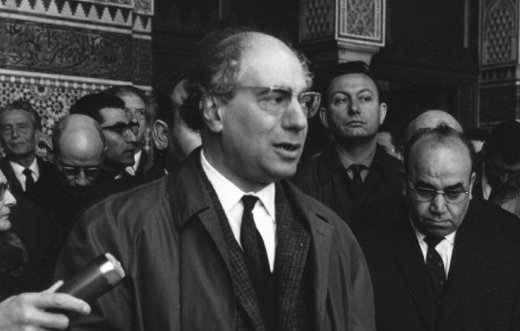 L'image montre un homme en train de prendre la parole devant un groupe de personnes. Il porte un manteau et des lunettes. Derrière lui, il y a plusieurs hommes et femmes qui l'écoutent attentivement. L'environnement semble formel, et il y a une atmosphère de rassemblement ou de cérémonie. En arrière-plan, on aperçoit des éléments d'architecture qui suggèrent un lieu historique ou important.