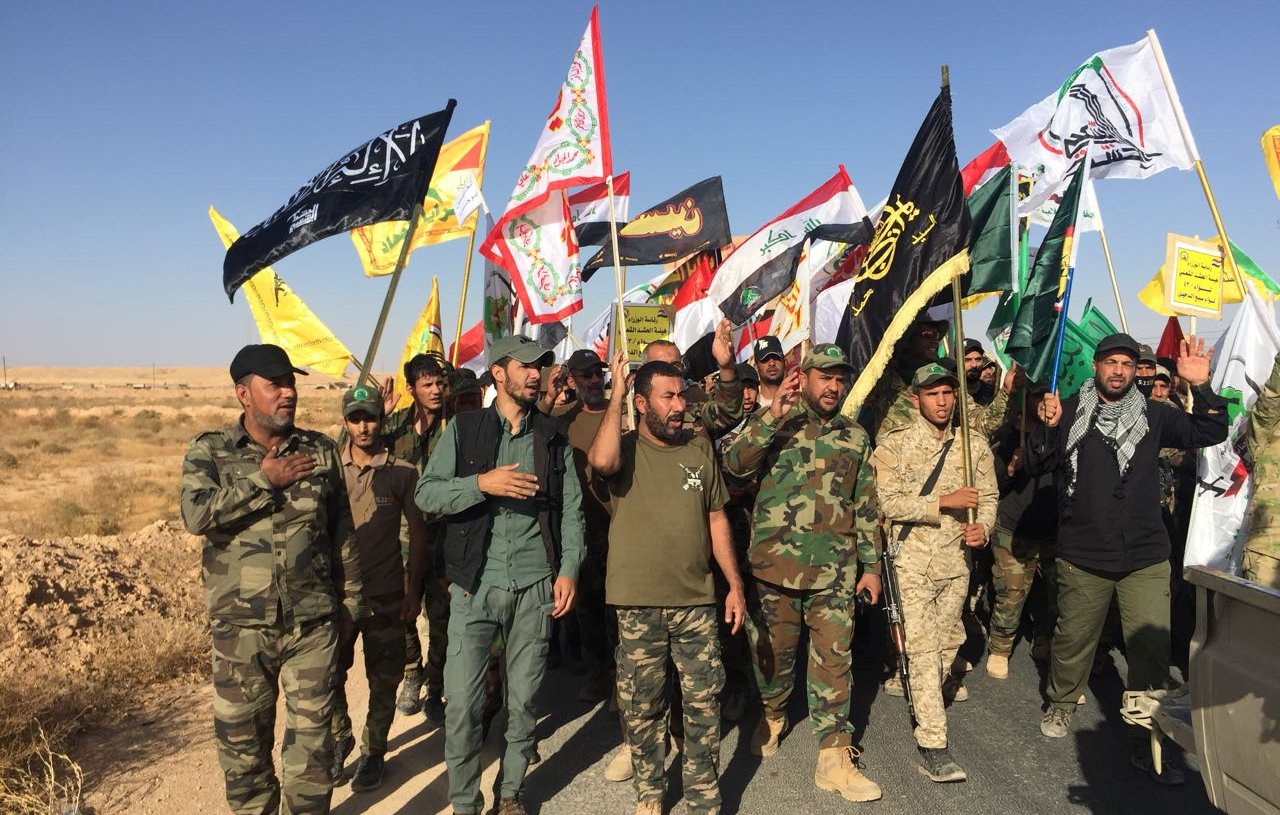 الصورة تظهر مجموعة من الأشخاص يسيرون معًا في مظاهرة أو مسيرة، يحملون أعلامًا متعددة الألوان. يبدو أنهم يرتدون ملابس عسكرية وبعضهم يرتدي زياً تقليدياً. الخلفية طبيعية، مع أراضٍ جافة وصحراوية. تعكس الصورة أجواء حماسية وتضامن بين المشاركين.