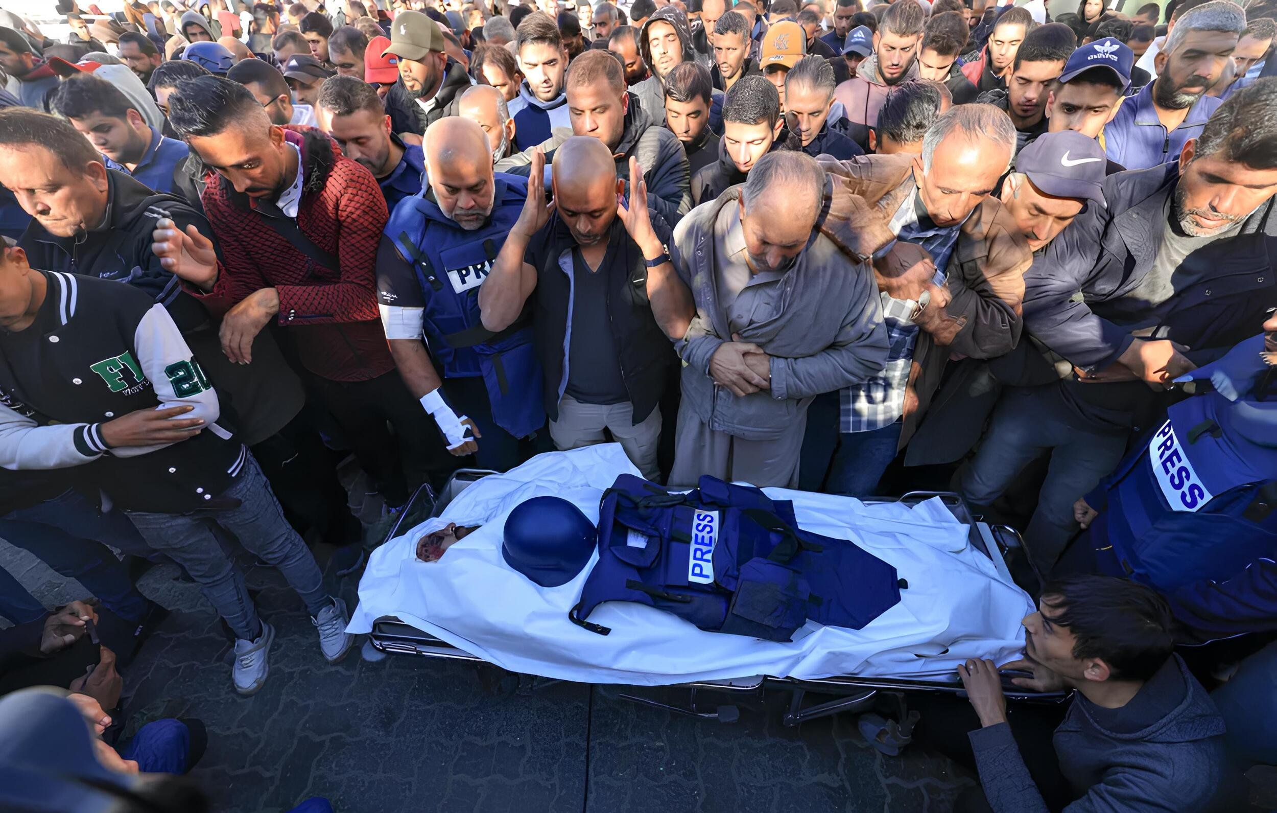 L'immagine mostra un gruppo di persone riunite in un'atmosfera di grande dolore e rispetto. Al centro, un corpo avvolto in un lenzuolo bianco è accompagnato da un giubbotto con la scritta "PRESS", suggerendo che si tratta di un giornalista. I presenti esprimono il loro cordoglio, alcuni con le mani giunte e altri con espressioni di tristezza. Il contesto sembra essere un evento commemorativo o funerario, evidenziando la gravità della situazione e il rispetto verso la persona deceduta.