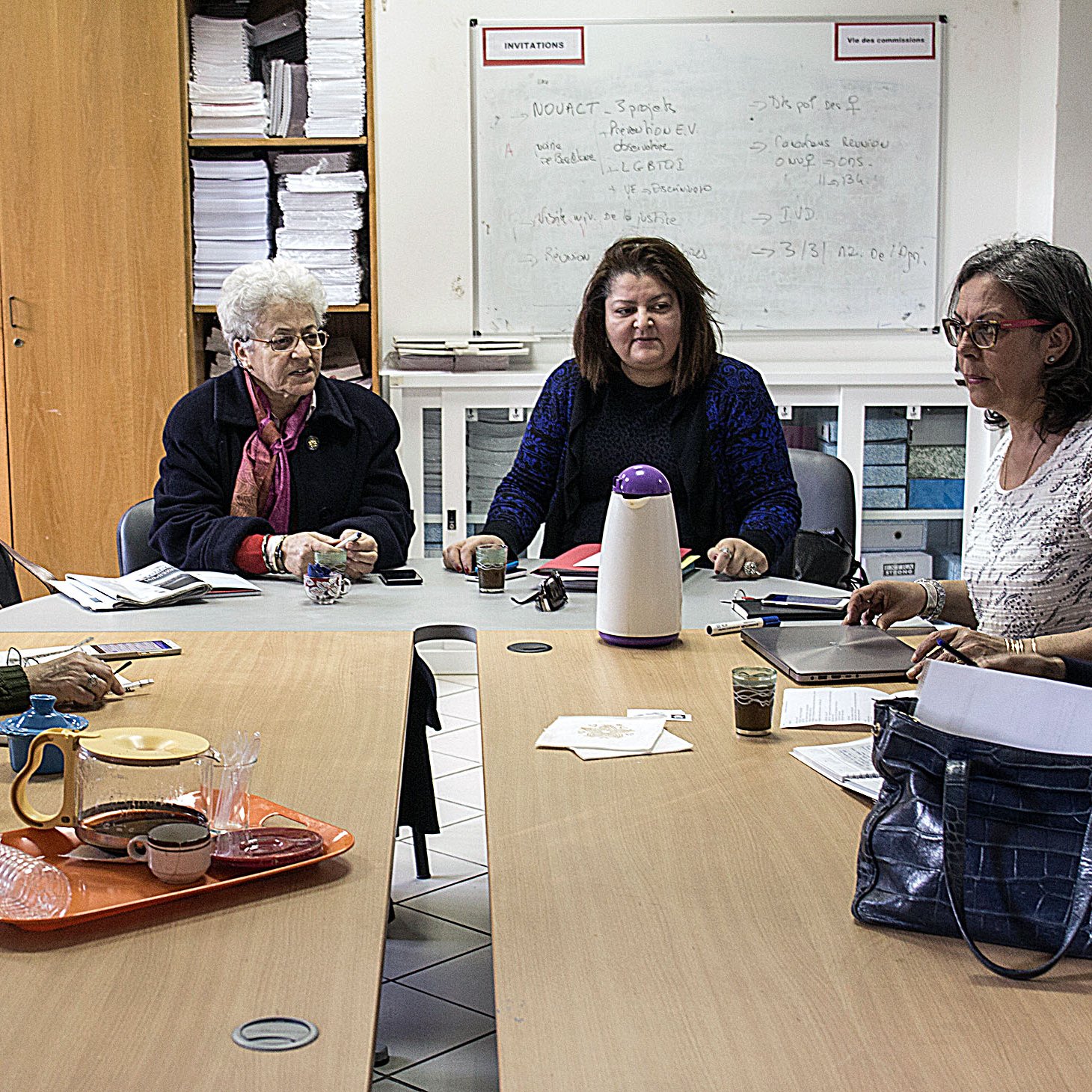 L'image montre un groupe de femmes assises autour d'une table dans une pièce qui semble être un espace de réunion. Elles sont engagées dans une discussion ou une activité collaborative. Sur la table, on peut voir des carnets, des verres et un thermos. L'environnement est simple, avec des étagères remplies de dossiers et de documents en arrière-plan. Les femmes semblent concentrées et impliquées dans leur échange.