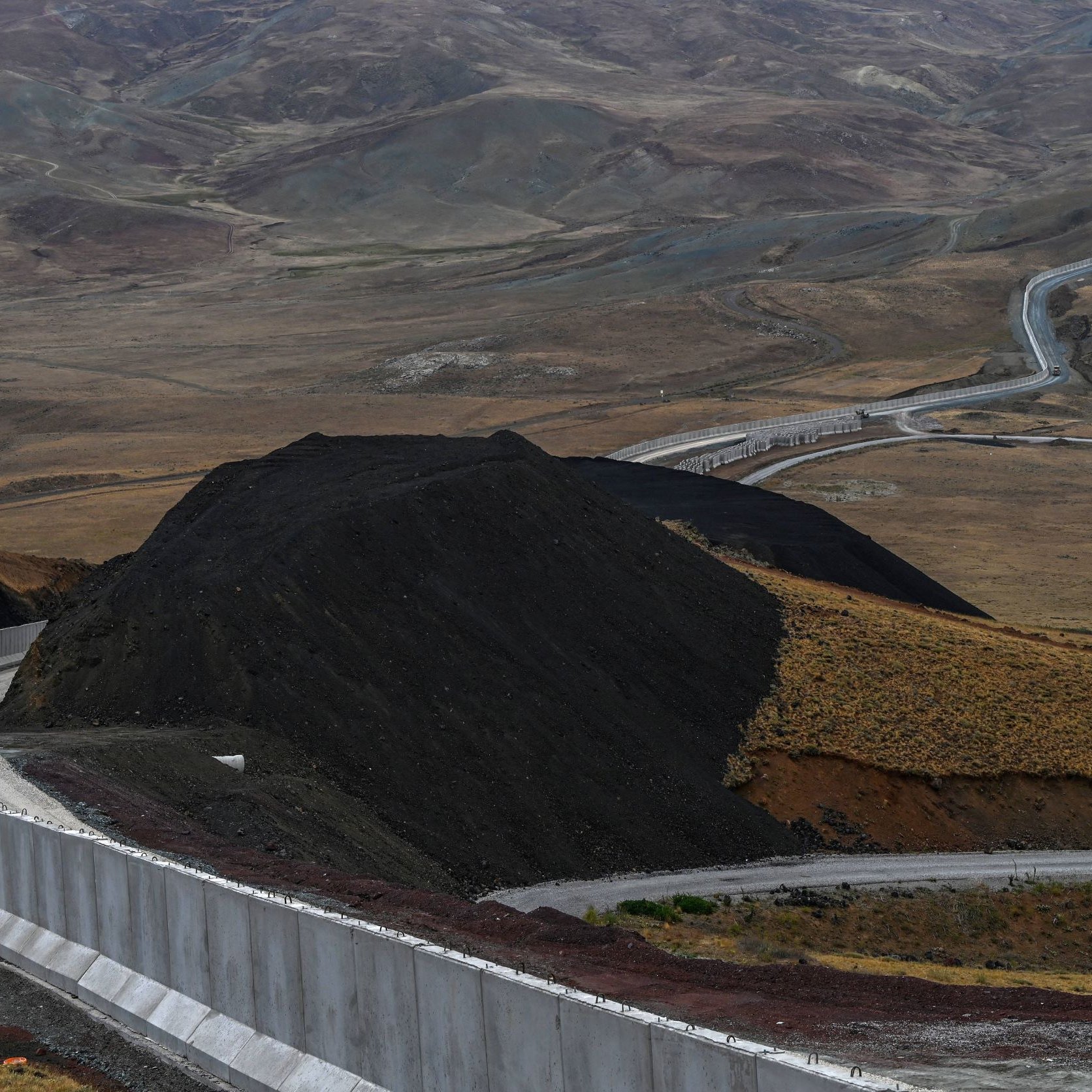 L'image montre un paysage vaste et aride, avec des collines et des montagnes en arrière-plan. Au premier plan, il y a un mur de béton qui serpente le long du sol, suivi d'une route qui s'étend dans la vallée. Le terrain est principalement composé de terre brune et de végétation clairsemée, avec des déclivités et des pentes. L'ambiance générale semble désertique et isolée.