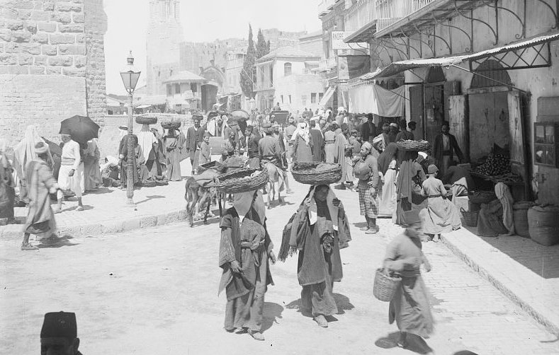 L'image montre une scène animée à la porte de Jaffa à Jérusalem. On y voit de nombreuses personnes en mouvement, portant des vêtements traditionnels. Certains portent des charges sur leur tête, tandis que d'autres se déplacent dans la rue. Des bâtiments historiques se dressent en arrière-plan, et l'atmosphère semble occuper un marché ou un lieu de rencontre. La lumière est vive, ce qui donne une impression de chaleur et d'activité.
