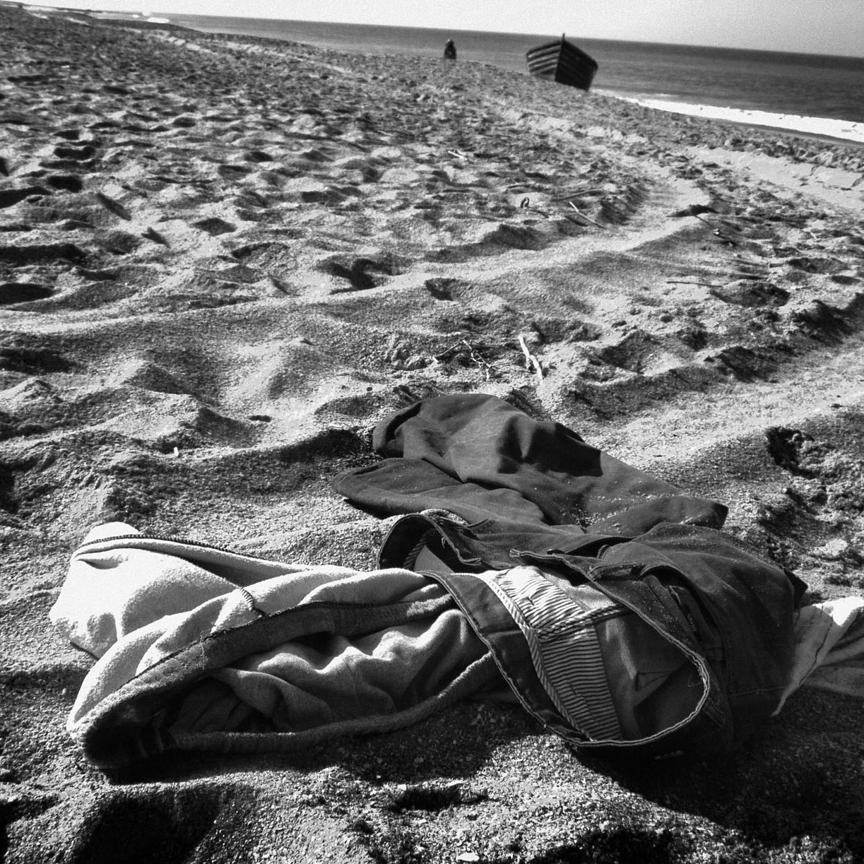 L'image montre un paysage de plage en noir et blanc, où des vêtements abandonnés sont éparpillés sur le sable. On peut voir des traces laissées par les pas dans le sable, créant un motif dans la texture. À l'arrière-plan, la mer se déroule tranquillement, et le ciel est clair. L'ensemble dégage une atmosphère de solitude et d'abandon.