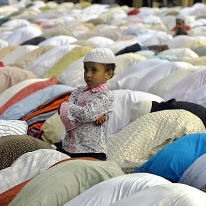 L'image montre un jeune garçon, vêtu d'une chemise à motifs et d'une petite kufi blanche, se tenant debout avec les bras croisés. Il regarde autour de lui, tandis qu'autour de lui, un grand nombre de personnes sont en prière, les corps inclinés et recouverts de vêtements colorés. L'ambiance semble empreinte de sérénité et de dévotion, avec des draps et des vêtements disposés sur le sol, typiques d'un rassemblement de prière.