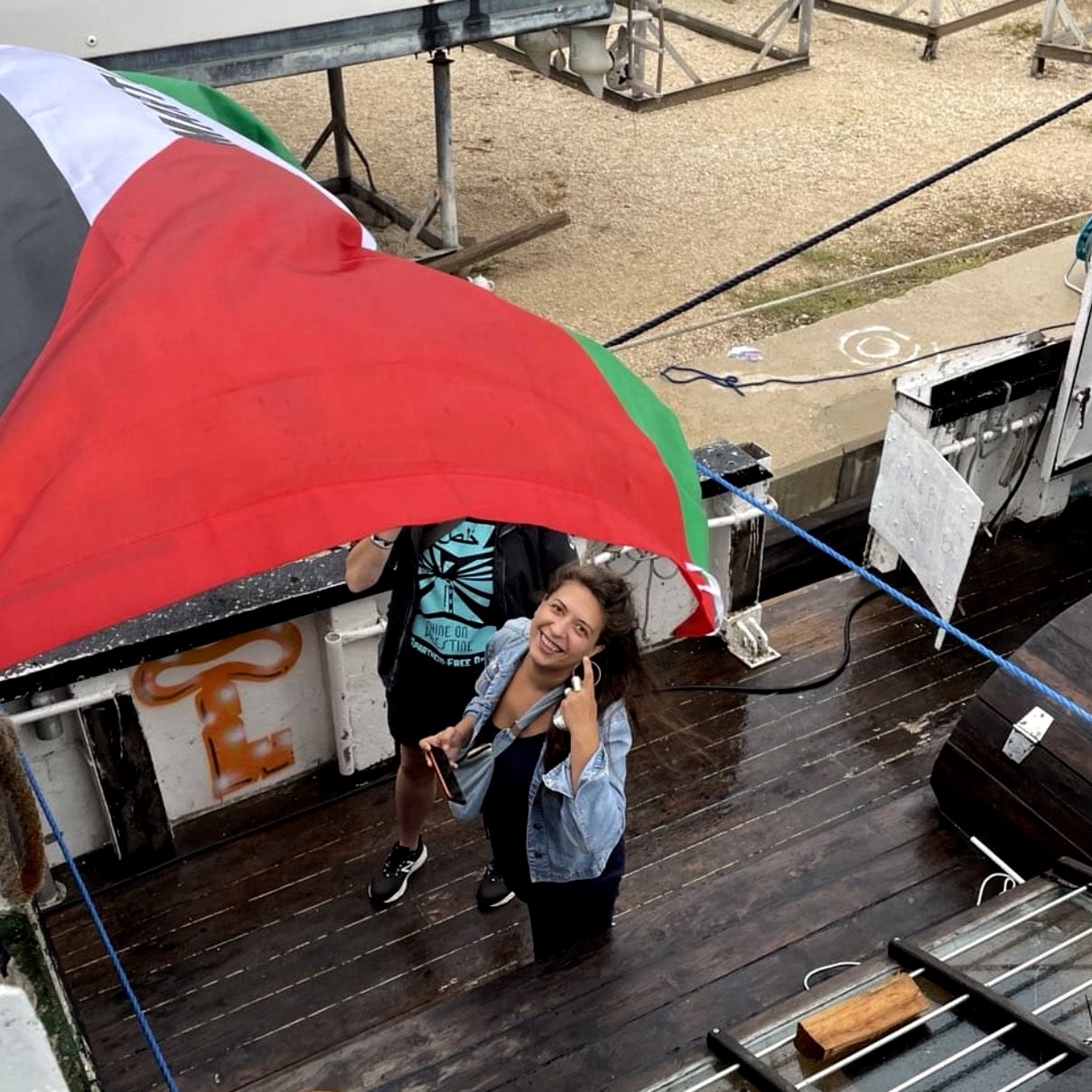 در این تصویر، دو نفر در حال کار با یک پرچم هستند که بر روی یک قایق یا سکوی چوبی قرار دارد. پرچم دارای رنگ‌های مشخصی است و یکی از افراد به دوربین لبخند می‌زند و انگشت شست خود را بالا برده است. محیط اطراف شامل تجهیزات قایق‌سواری و فضای باز است. به نظر می‌رسد این افراد در حال آماده‌سازی یا نصب پرچم هستند.