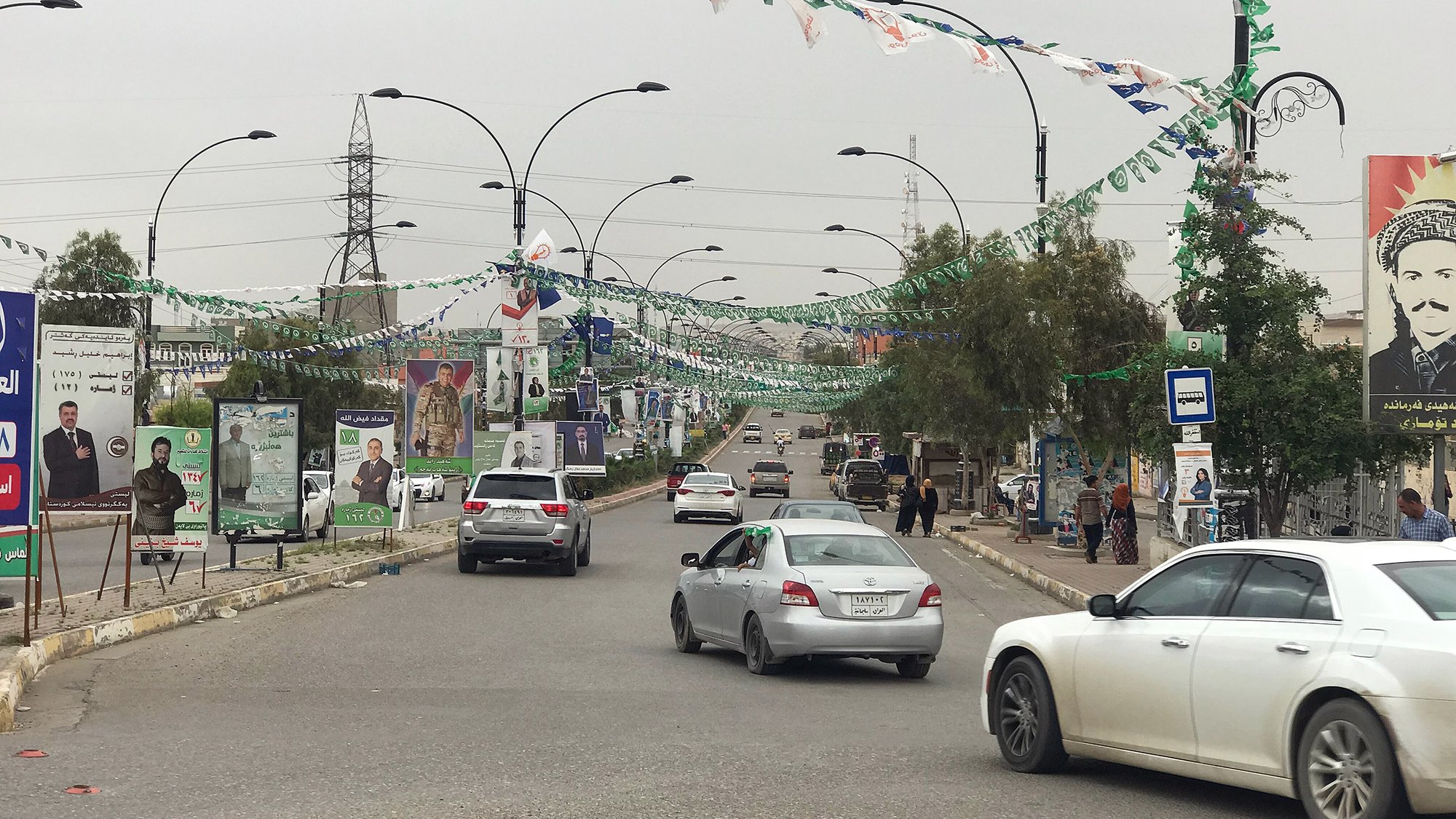 L'image montre une rue bordée de drapeaux et de bannières, probablement en préparation d'un événement ou d'une célébration. On peut voir plusieurs voitures circulant sur la route. À côté de la route, il y a des panneaux avec des affiches, ce qui indique une zone urbaine active. Des personnes se trouvent également sur le trottoir, et des arbres et des lampadaires sont présents le long de la rue, donnant un aspect dynamique à la scène. Le ciel semble nuageux, suggérant une ambiance calme.