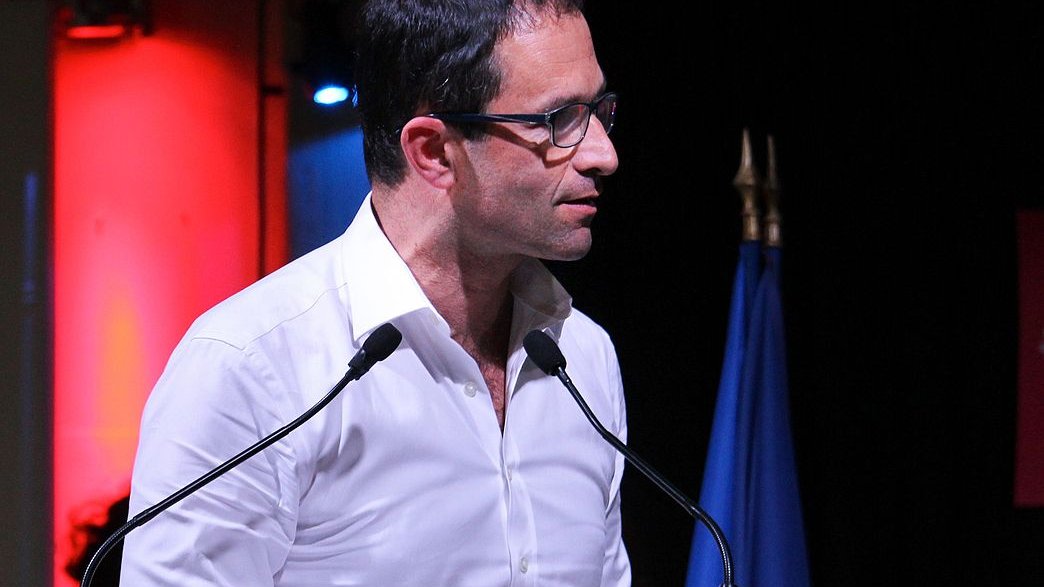 L'image montre un homme debout, parlant à un podium avec le nom "Benoît Hamon 2017" affiché sur le devant. Il porte une chemise blanche et des lunettes, et il semble s'exprimer avec passion. En arrière-plan, on peut voir des éclairages colorés, notamment en rouge, et un drapeau français. L'homme semble engagé dans un discours ou une présentation.