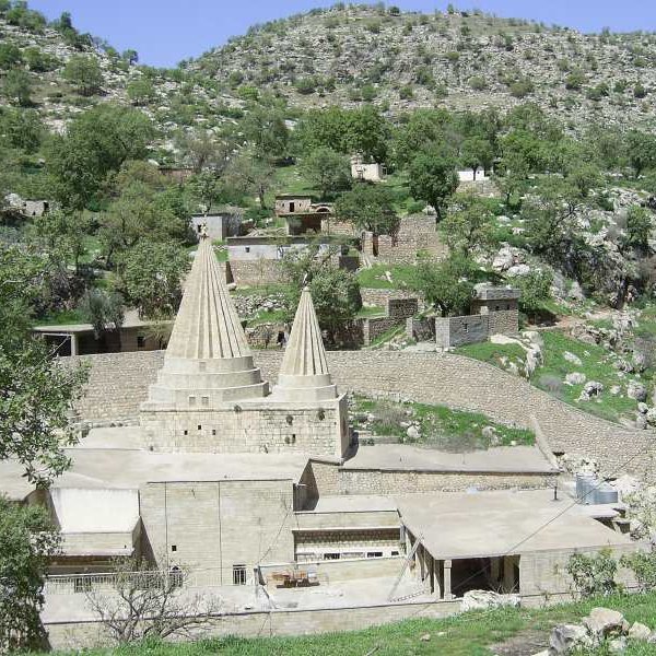 L'image montre un paysage montagneux avec une structure architecturale distinctive au centre. Cette structure présente deux tours ou pinacles coniques, typiques d'un style architectural particulier. Autour, on aperçoit des bâtiments en pierre et en béton, ainsi que des arbres verdoyants qui ajoutent une touche de nature. Le terrain est légèrement accidenté, avec des collines à l'arrière-plan, créant un environnement pittoresque et tranquille.