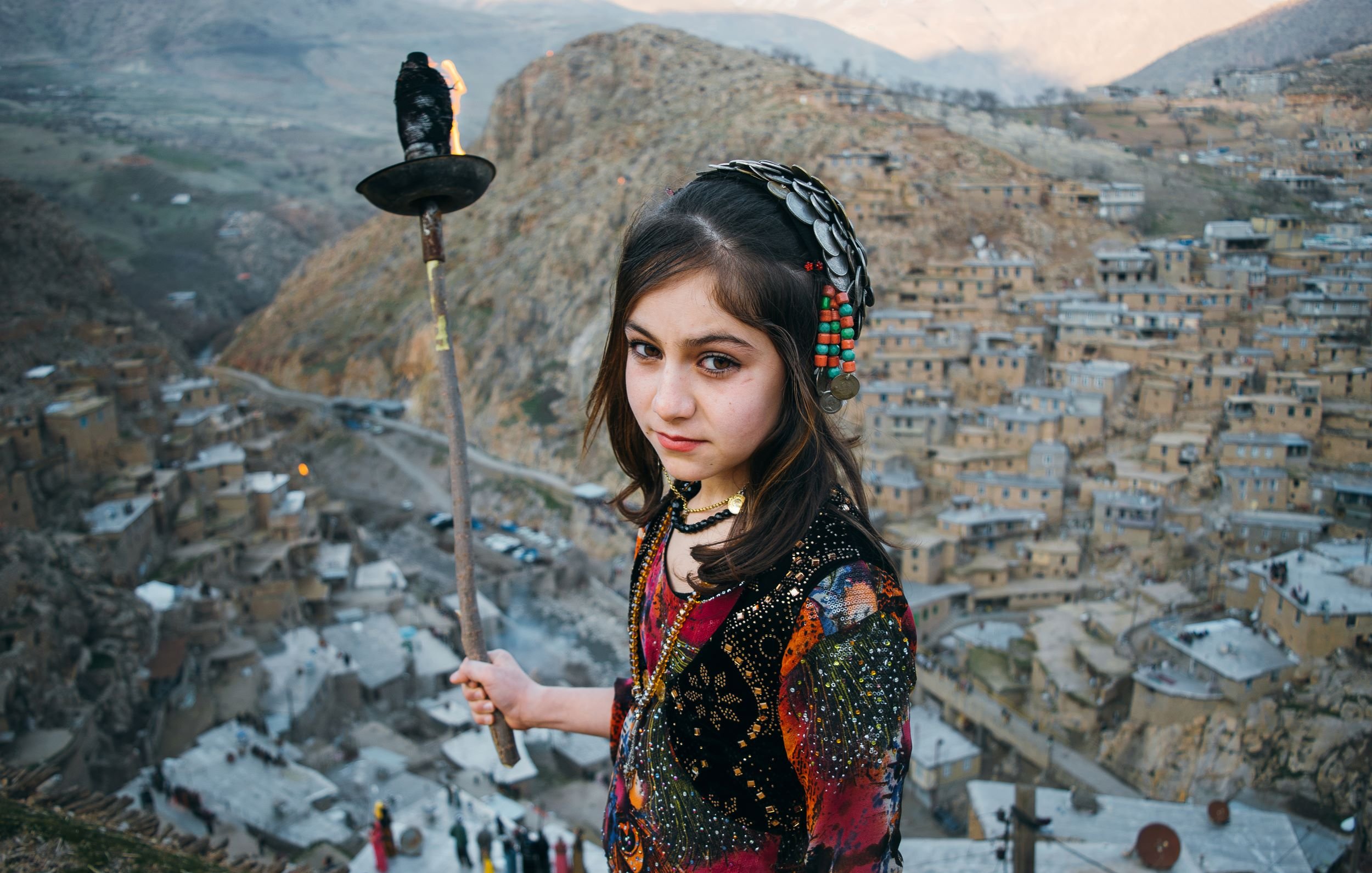 L'image montre une jeune fille tenant une torche, se tenant au sommet d'une colline surplombant un village pittoresque. Elle porte des vêtements colorés ornés de motifs traditionnels, et son regard est à la fois sérieux et perçant. En arrière-plan, on aperçoit des maisons construites à flanc de montagne, avec des paysages verdoyants s'étendant au loin. L'atmosphère de la scène est à la fois authentique et captivante, évoquant des traditions anciennes et une culture riche.