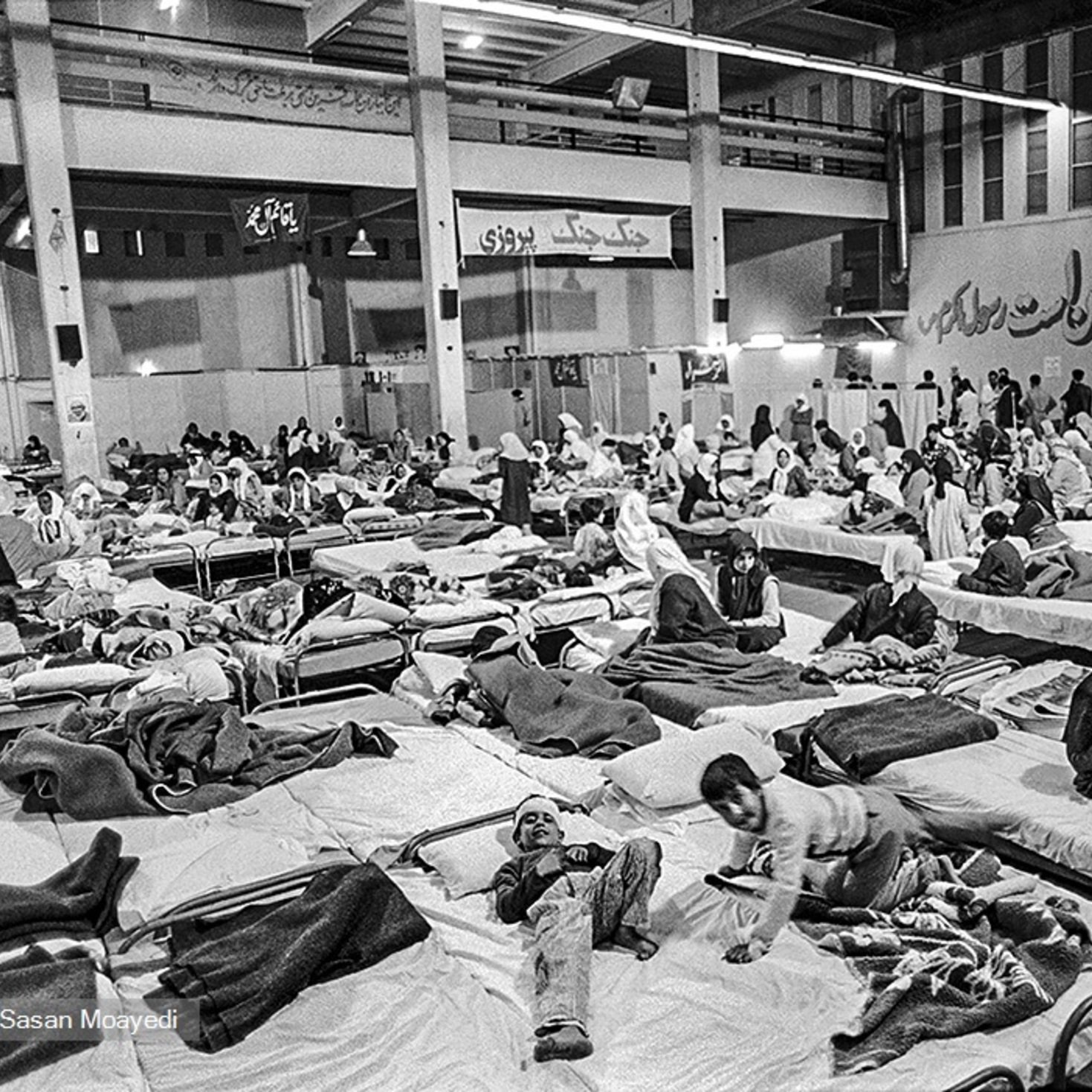 این تصویر یک فضای داخلی را نشان می‌دهد که پر از تخت‌های بیمارستانی است. افراد مختلفی در حالت‌های مختلف دراز کشیده‌اند، برخی در حال استراحت و برخی دیگر در حال حرکت یا صحبت با یکدیگر هستند. محیط به نظر شلوغ و پر از فعالیت می‌رسد، و پوشش بیشتر افراد ساده و شامل چادر یا پتو است. دیوارها به نوعی دکور شده‌اند، و فضای کلی شاید به یک مرکز درمانی یا پناهگاه اشاره دارد.