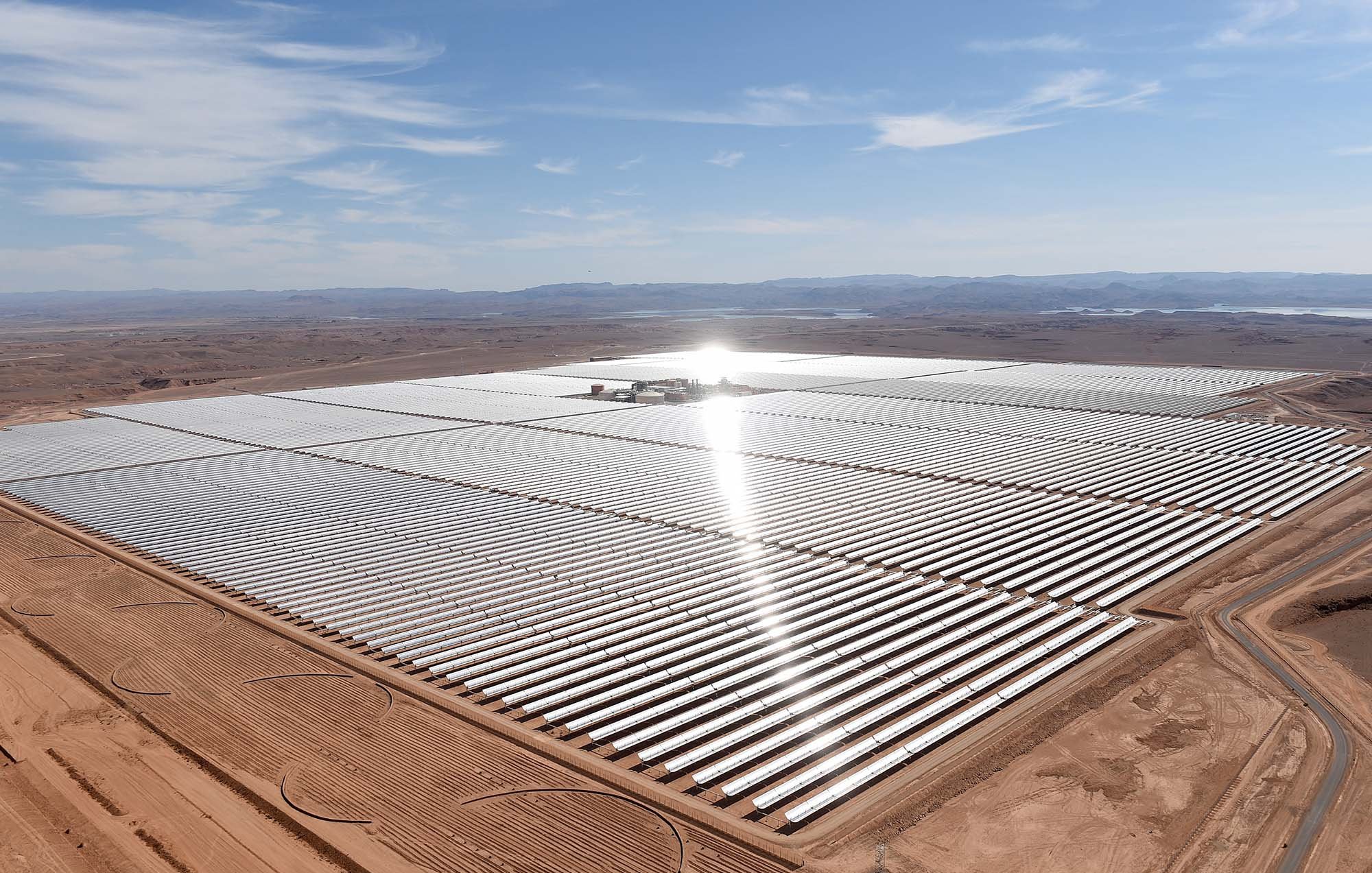 L'image montre une vaste installation de panneaux solaires, s'étendant sur une grande surface de terrain désertique. Les panneaux sont disposés en rangées régulières, reflétant la lumière du soleil. On peut apercevoir des collines à l'arrière-plan, et le ciel est clair avec quelques nuages. L'ensemble donne une impression de modernité et de durabilité, soulignant l'importance des énergies renouvelables dans le paysage contemporain.