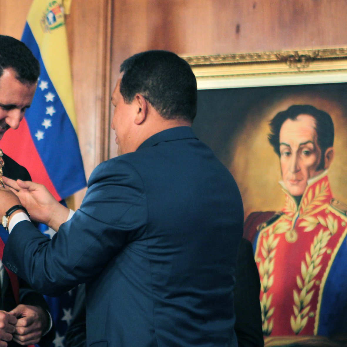 L'image montre une cérémonie officielle où un homme, probablement un leader politique, reçoit un insigne ou un collier de la part d'un autre homme. Le premier homme est vêtu d'un costume noir et porte une écharpe, tandis que le second, habillé en bleu, est en train de lui remettre l'insigne autour du cou. En arrière-plan, on aperçoit un portrait d'une figure historique, ainsi que des drapeaux, ce qui suggère un contexte politique et national.