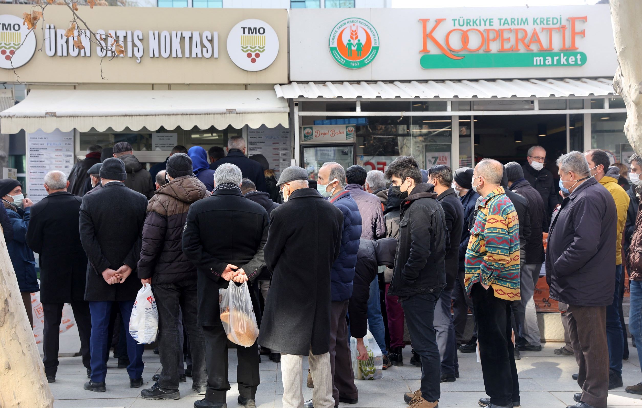 L'image montre une foule de personnes faisant la queue devant un magasin, probablement un marché ou un point de vente. Les gens semblent attendre pour entrer ou acheter des produits. Ils portent des vêtements d'hiver, et certains tiennent des sacs. L'atmosphère semble calme, mais il y a une certaine impatience dans la file d'attente. Au-dessus de l'entrée du magasin, on peut voir des panneaux indiquant le nom et la fonction de l'établissement.