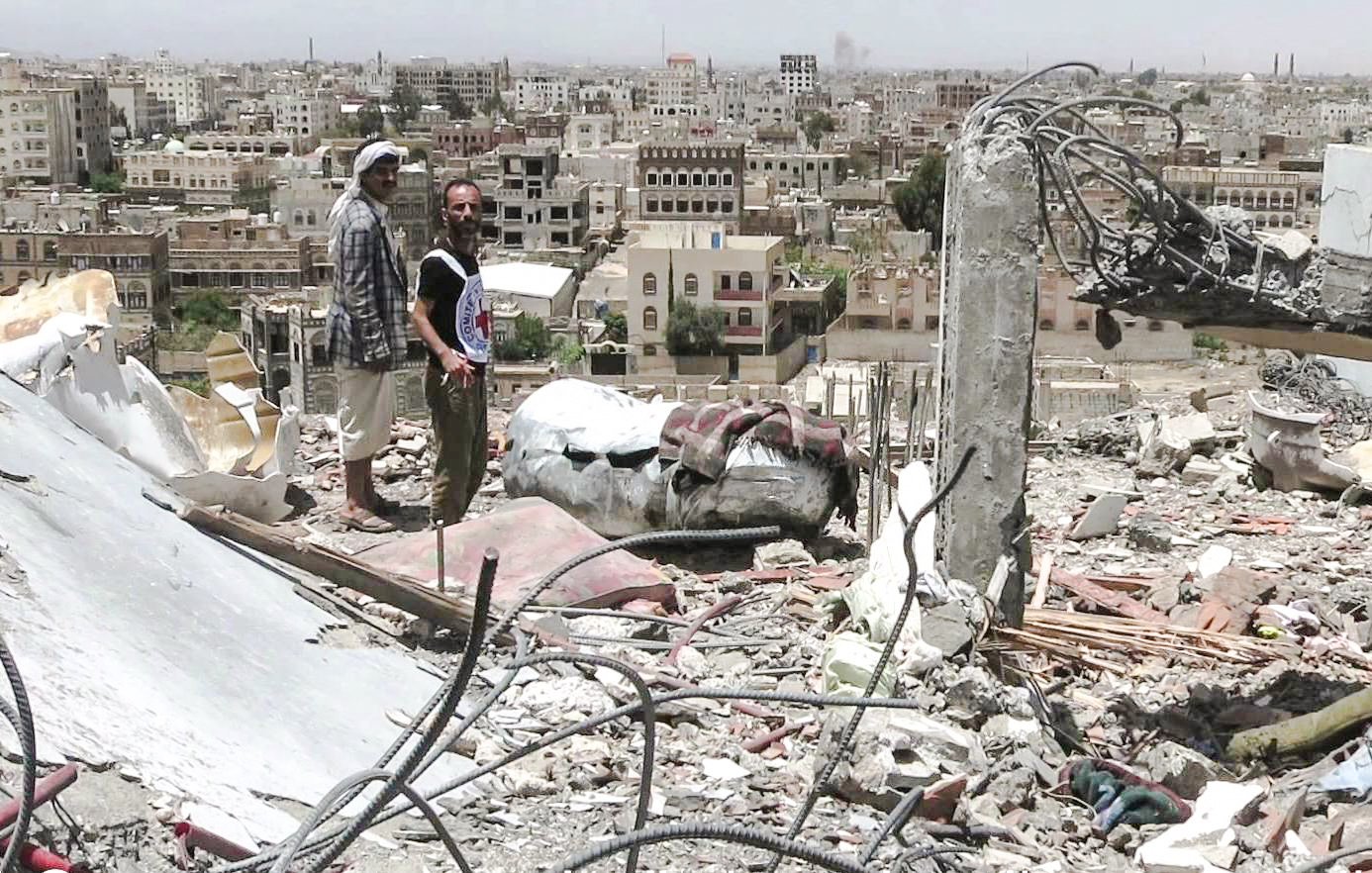 تظهر الصورة مشهدًا من مدينة مدمرة، حيث يقف رجلان على أنقاض مبنى. خلفهما، ترتفع مجموعة من المباني في المدينة، وهي تبدو متضررة بشكل كبير. يبدو أن البيئة المحيطة تعكس آثار الدمار، مع الحطام والمخلفات المنتشرة في كل مكان. السماء تبدو مشمسة، مما يبرز الف contrast بين الدمار والبيئة المحيطة.