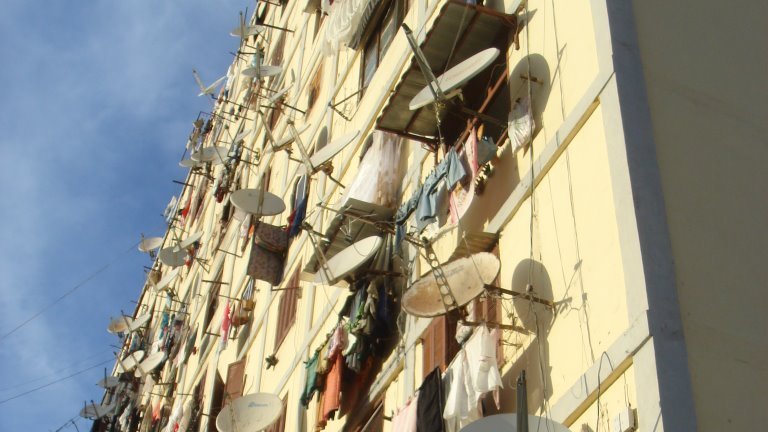 L'image montre un bâtiment d'habitation avec de nombreux balcons. Les murs de l'immeuble sont couverts de vêtements suspendus à sécher, créant un décor coloré et animé. De plus, plusieurs antennes parabolique sont fixées sur les façades, indiquant que les habitants ont accès à la télévision satellite. Le ciel est clair, ajoutant une lumière naturelle à la scène. L'ensemble donne une impression de vie communautaire et de simplicité dans un cadre urbain.