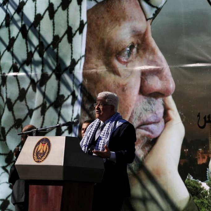 L'image représente un homme en train de parler lors d'un discours. Il se tient devant un grand poster qui affiche le portrait d'un homme en keffieh, regardant pensivement au loin. Le décor suggère qu'il s'agit d'un événement commémoratif, probablement lié à des thèmes politiques ou historiques. Le podium sur lequel l'homme s'exprime est orné d'un emblème officiel, tandis que le fond montre une ville, ajoutant un aspect symbolique à la scène.