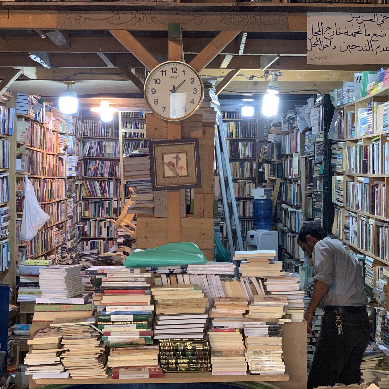 L'image montre une librairie pleine de livres, avec des étagères qui montent jusqu'au plafond. Il y a une grande horloge murale qui indique l'heure. Au centre, des piles de livres sont empilées sur une table, tandis qu'un homme est penché sur une étagère, en train de chercher ou de réorganiser des livres. L'ambiance semble chaleureuse et accueillante, avec un éclairage doux provenant de lampes suspendues. Une inscription en arabe est visible au mur.