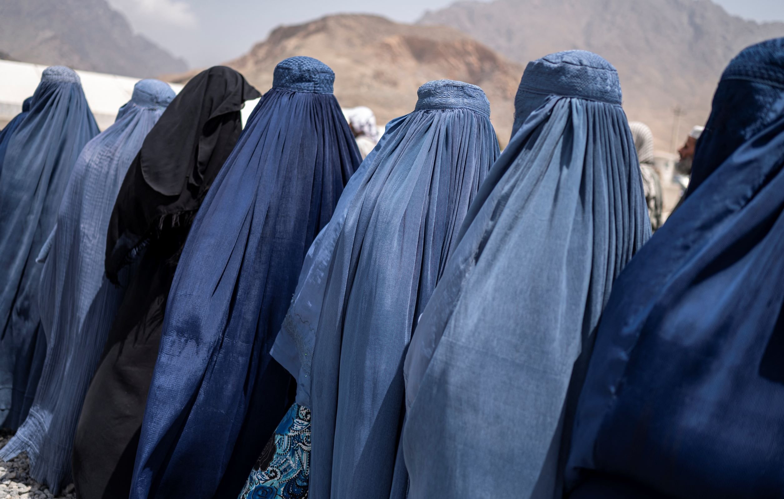 L'image montre un groupe de femmes de dos, vêtues de burqas de différentes nuances de bleu, se tenant en ligne. On peut également apercevoir une femme portant des vêtements noirs. En arrière-plan, on distingue des montagnes, et le ciel semble nuageux. L'ambiance générale semble être celle d'un rassemblement ou d'une rencontre.