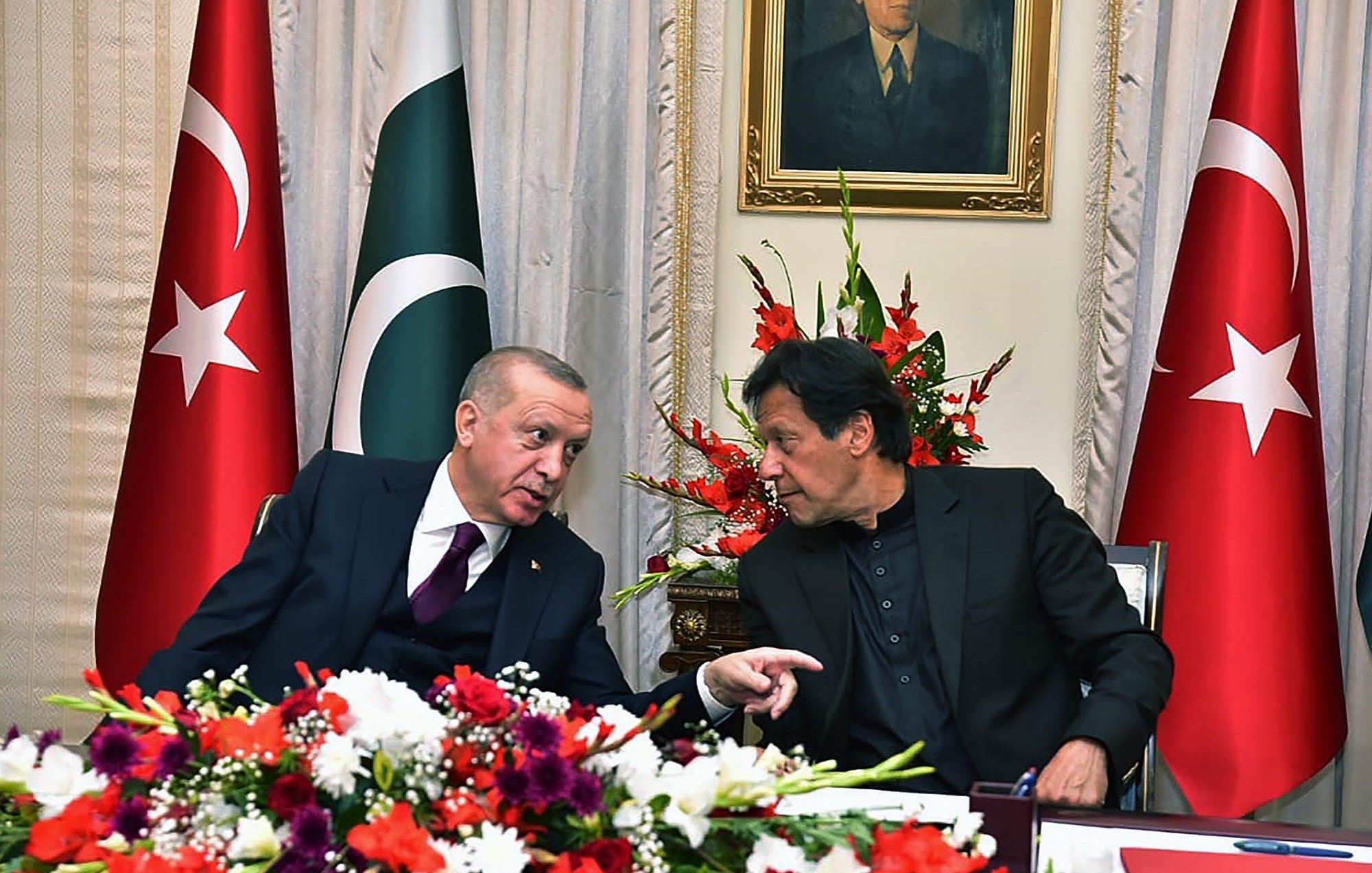 L'image montre deux hommes en conversation, assis à une table décorée de fleurs. En arrière-plan, on peut voir des drapeaux turc et pakistanais, ainsi qu'un portrait accroché au mur. Les hommes semblent échanger des idées de manière animée, et l'atmosphère semble conviviale.