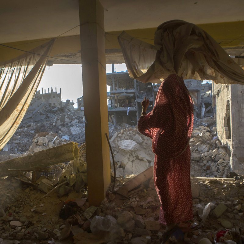 L'image montre une scène à l'intérieur d'un bâtiment endommagé. On aperçoit une femme vêtue d'un vêtement traditionnel, se tenant près d'une fenêtre qui donne sur des ruines. De l'autre côté, on voit des décombres de bâtiments effondrés, laissant entrevoir la désolation d'un environnement détruit. La lumière du soleil filtre à travers les rideaux, ajoutant une ambiance à la fois triste et poignante à la scène.