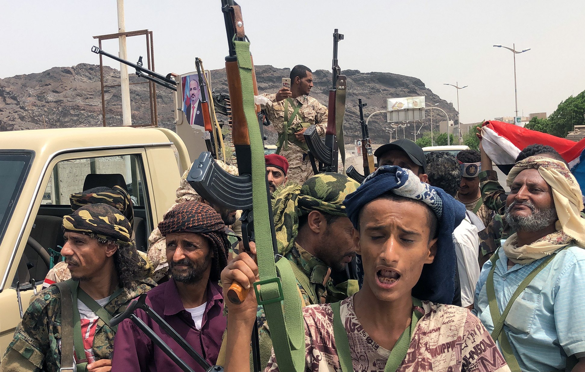 در این تصویر گروهی از مردان مسلح در یک منطقه عمومی دیده می‌شوند. برخی از آن‌ها کلاهی به سر دارند و در دست سلاح‌هایی نظیر کلاشنیکف نگه داشته‌اند. در پس‌زمینه، تصاویر و پرچم‌هایی از یمن قابل مشاهده است. به نظر می‌رسد آن‌ها در حال برگزاری یک گردهمایی یا نمایش نیرو هستند. زمینه کوهستانی و ساختمانی در دوردست قرار دارد. جایگاه‌های زیادی از سوی خودروها در اطراف آن‌ها وجود دارد.