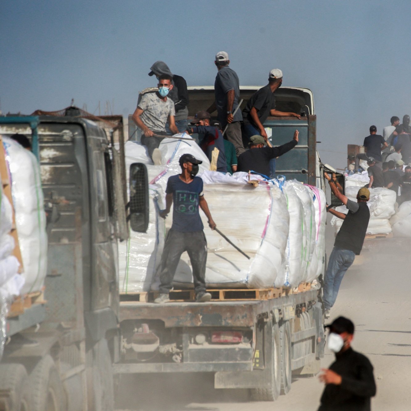 تصویر شامل کامیون‌هایی است که بار سنگینی حمل می‌کنند و عده‌ای در حال کار در یک محیط غبارآلود هستند.