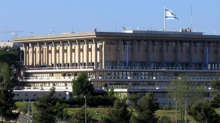 L'image montre un bâtiment moderne avec une architecture distincte, probablement situé dans un environnement urbain. Le bâtiment a plusieurs niveaux, avec des fenêtres et des balcons visibles. On peut également apercevoir des drapeaux, y compris celui d'Israël, flottant en haut du bâtiment. Autour, il y a des arbres et de la verdure, ce qui donne une impression de nature intégrée dans un cadre urbain. Le ciel est bleu et dégagé, ajoutant à la luminosité de la scène.