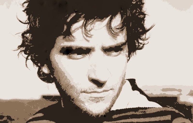 L'image présente un homme avec des cheveux bouclés et une expression sérieuse. Son visage est éclairé d'une manière qui met en valeur ses traits, et il semble regarder au loin. Le fond est flou, ce qui met l'accent sur son visage. La palette de couleurs est principalement en nuances sépia, ce qui donne une atmosphère nostalgique ou intemporelle à l'image.