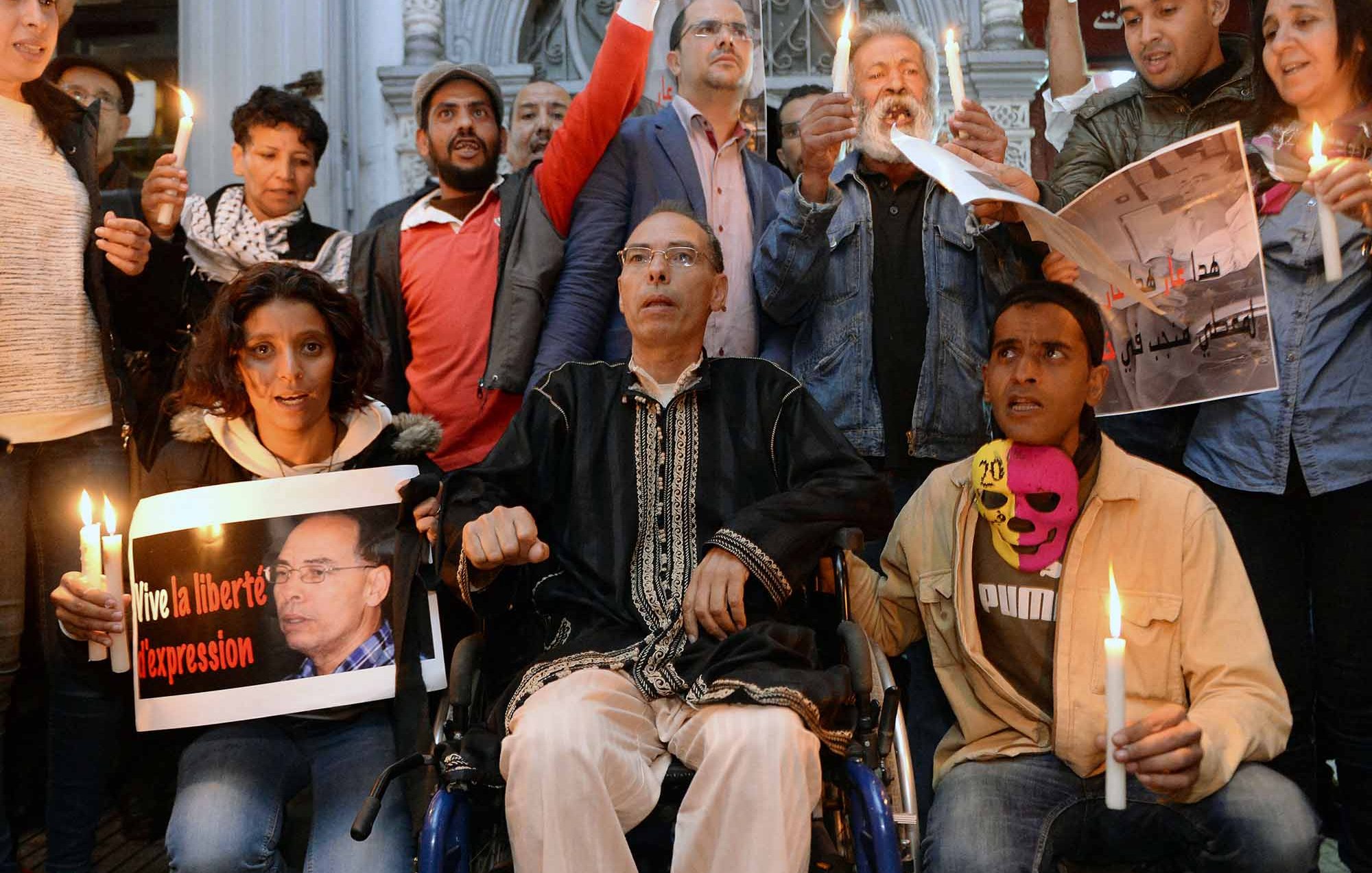 L'image montre un groupe de personnes rassemblées, tenant des bougies allumées. Au premier plan, un homme en fauteuil roulant est au centre, tenant une pancarte. Les autres personnes autour de lui semblent participer à une manifestation pacifique, certaines brandissant des affiches et d'autres affichant des expressions sérieuses. L'ambiance semblerait être axée sur la défense des droits et de la liberté d'expression.