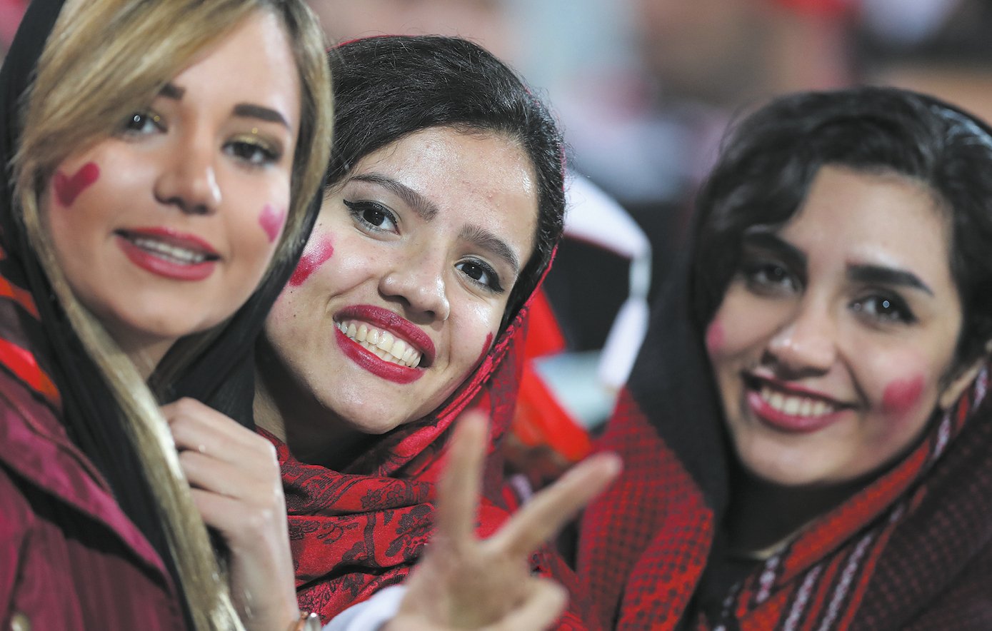 L'image montre trois femmes souriantes, vêtues de vêtements dans des tons sombres, avec des écharpes ou des foulards sur la tête. Chacune d'elles a des cœurs rouges dessinés sur les joues, ce qui suggère qu'elles célébrent un événement sportif ou une occasion festive. Elles affichent des expressions de joie et de camaraderie, et l'une des femmes montre un signe de paix avec ses doigts. L'arrière-plan est flou, mais on peut discerner une ambiance festive.