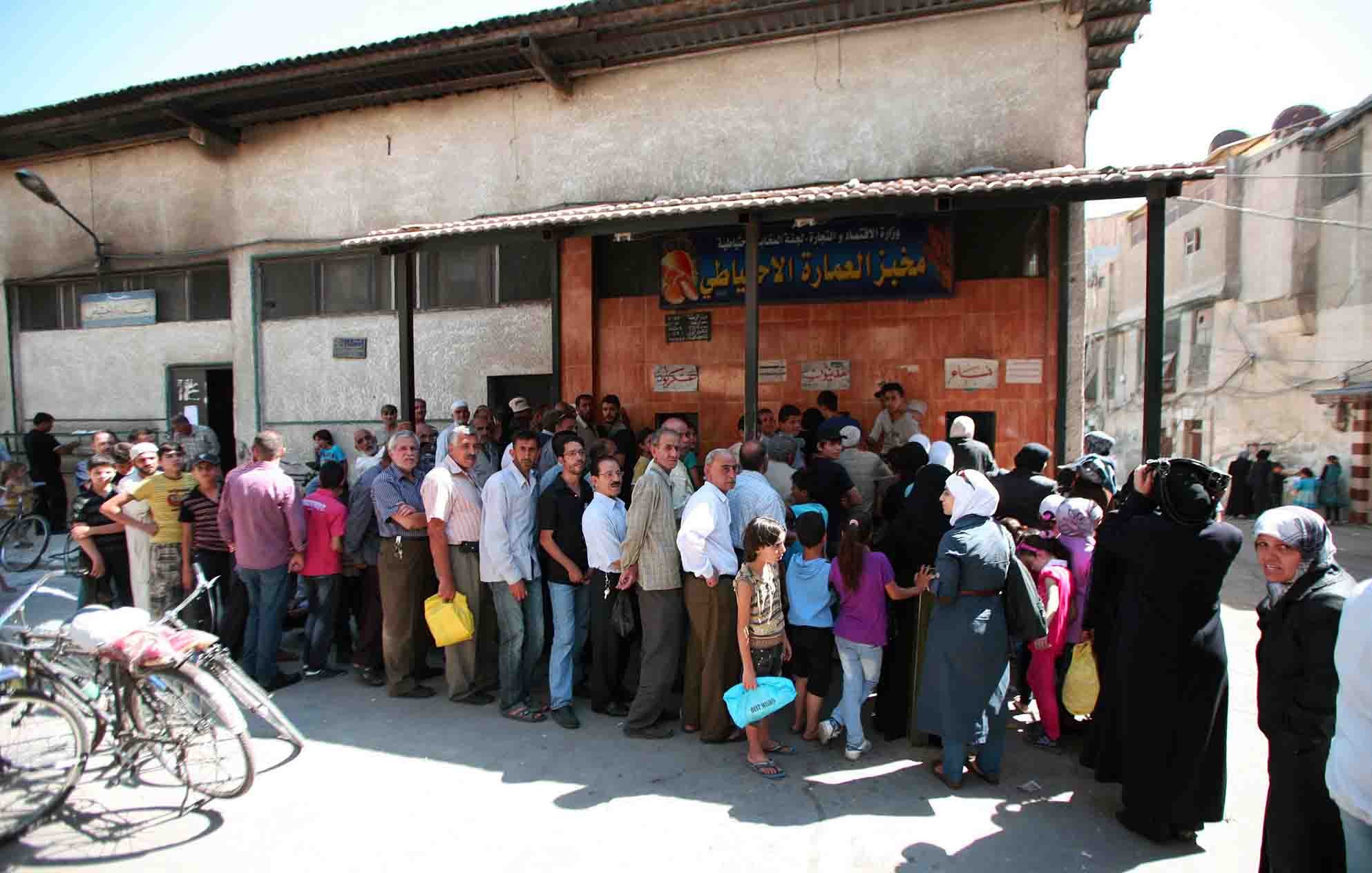 L'immagine mostra una lunga fila di persone che si trovano all'esterno di un edificio. La scena sembra essere in un contesto urbano, probabilmente in una città del Medio Oriente. Le persone nella fila sono di diverse età e indossano abiti vari, alcuni dei quali tradizionali. Alcuni individui portano sacche o borse, e si può notare un'atmosfera di attesa e di comunità. Sullo sfondo, ci sono edifici semplici con elementi architettonici tipici della regione.