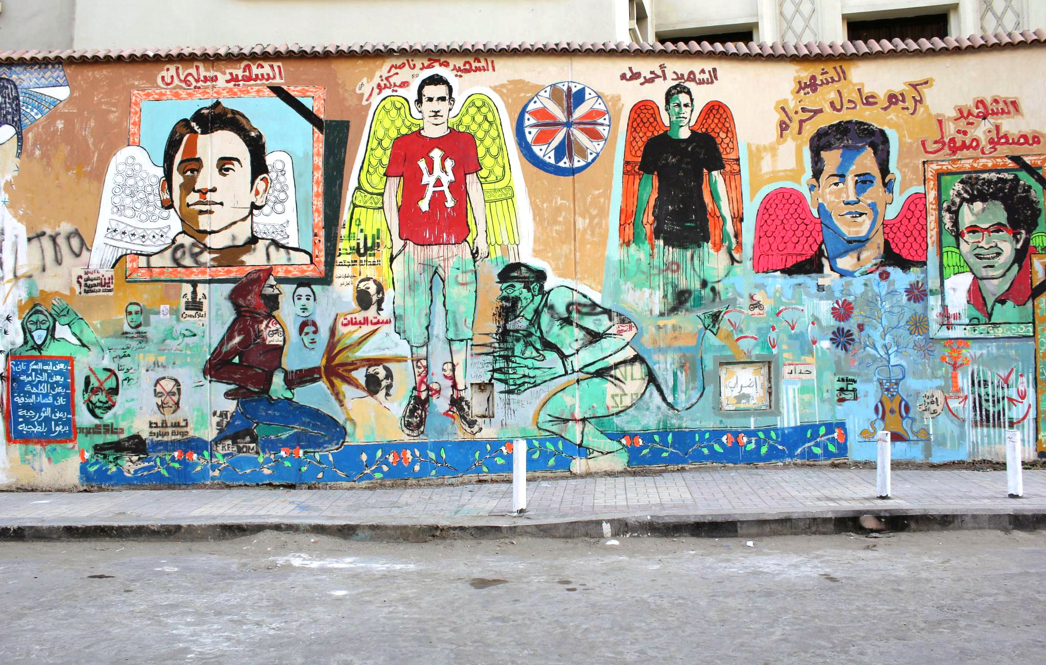 L'image représente une fresque murale colorée sur un mur. On y voit plusieurs portraits d'individus, accompagnés de motifs d'ailes et de détails symboliques. Les personnages ont des expressions variées, et des écritures en arabe sont visibles en haut de la fresque. Le fond présente des éléments artistiques tels que des fleurs et d'autres motifs graphiques. L'ambiance générale est vibrante et évoque un message fort, probablement en lien avec la culture locale ou un hommage à des figures importantes. Le sol devant le mur est en béton, et on peut y voir des supports en métal.