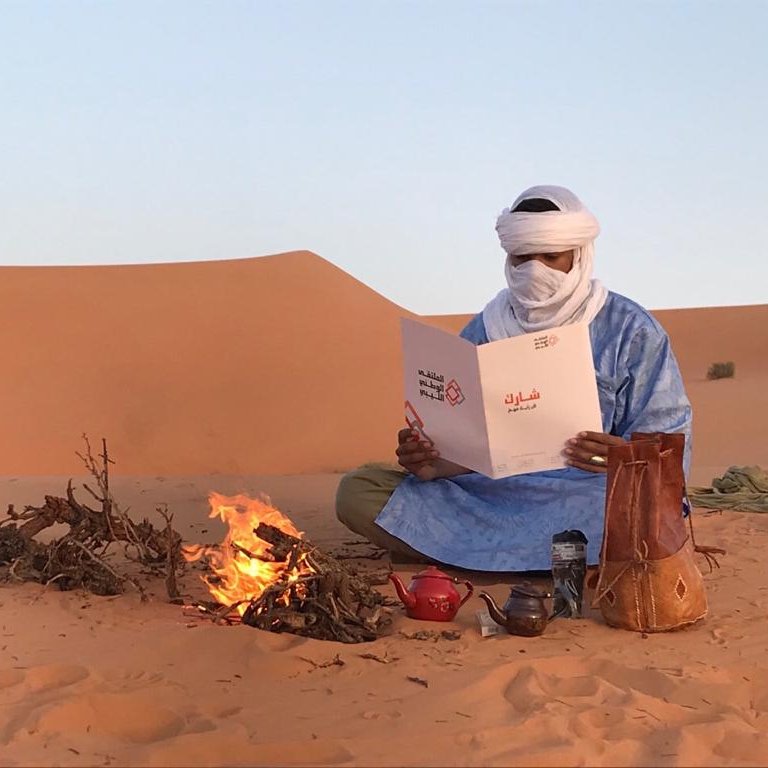 L'image montre un homme assis dans le désert, portant des vêtements traditionnels bleus et un turban. Il est devant un petit feu de camp, où des brindilles brûlent. L'homme tient un menu qu'il examine attentivement. À côté de lui, il y a une théière et un pot en terre cuite. En arrière-plan, on peut voir des dunes de sable sous un ciel clair. L'ambiance évoque la tranquillité et la culture nomade.