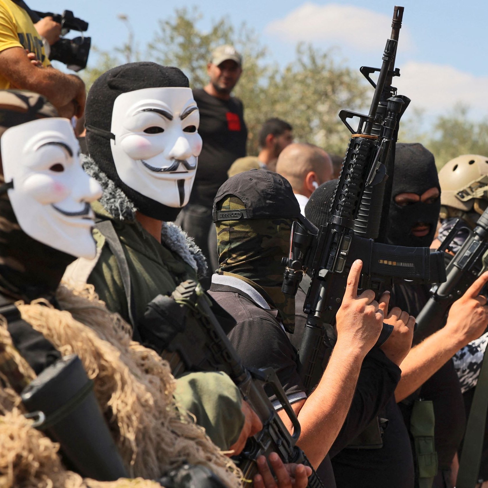 L'image montre un groupe de personnes portant des masques et des vêtements militaires, tenant des fusils. Les visages des individus sont principalement cachés, et ils semblent formés en ligne, chacun montrant une arme. En arrière-plan, on aperçoit d'autres personnes observant la scène. L'atmosphère semble tendue et engagée.