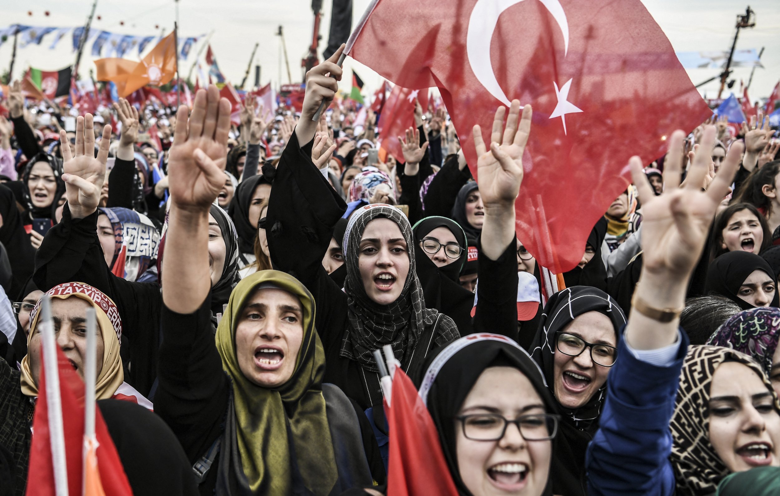 الصورة تظهر حشودًا من النساء في تظاهرة أو فعالية جماهيرية. العديد من النساء يحملن الأعلام التركية ويعبرن عن حماسهن من خلال رفع الأيادي. يبدو أنهن يرتدين ملابس تقليدية أو حجاب، مما يعكس تنوع أساليب التعبير عن الهوية. الجو العام يظهر حيوية وشغفًا، مع وجود لافتات وأعلام في الخلفية تدل على رسالة أو هدف مشترك.