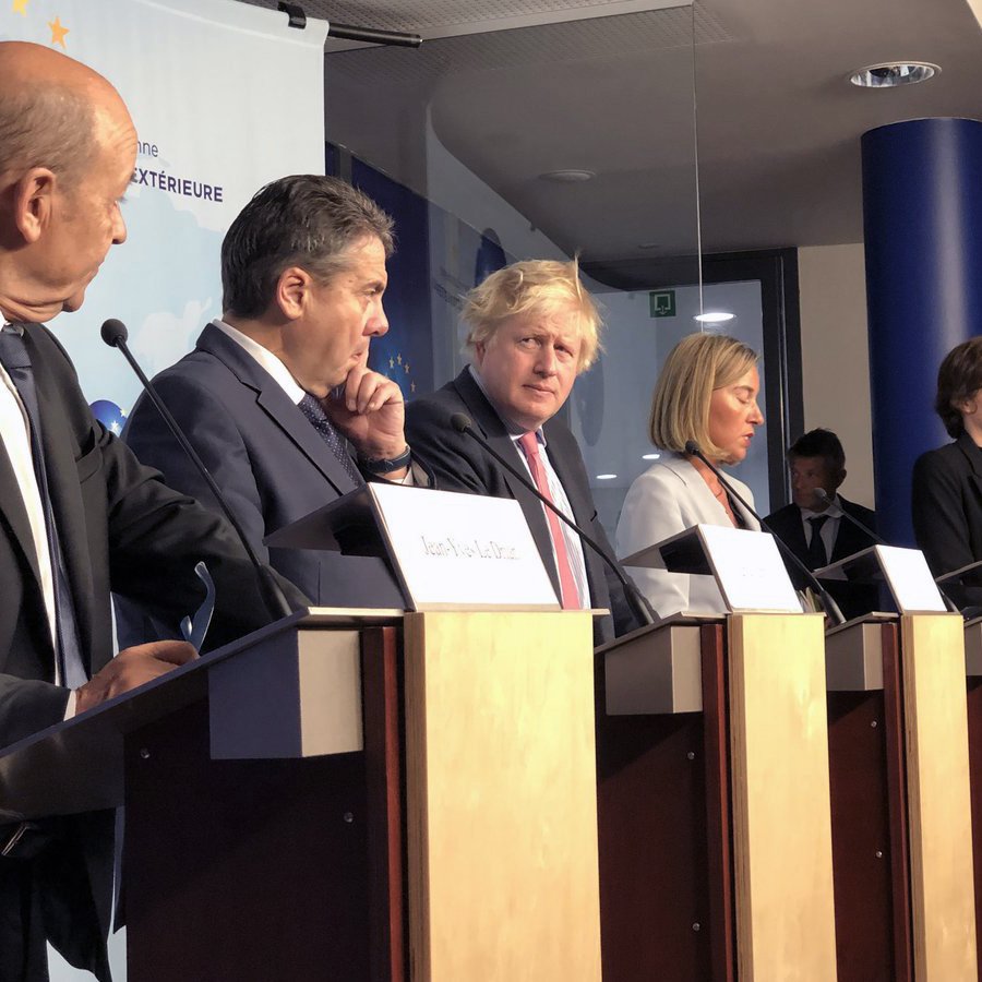 L'image montre une conférence de presse avec plusieurs personnes debout derrière des pupitres. Parmi eux, on peut voir des hommes en costume et une femme en tenue formelle. Ils semblent être en train de discuter ou d'écouter des questions, avec une atmosphère sérieuse. L'arrière-plan présente des éléments visuels représentant des thèmes internationaux, comme des logos ou des symboles géographiques.