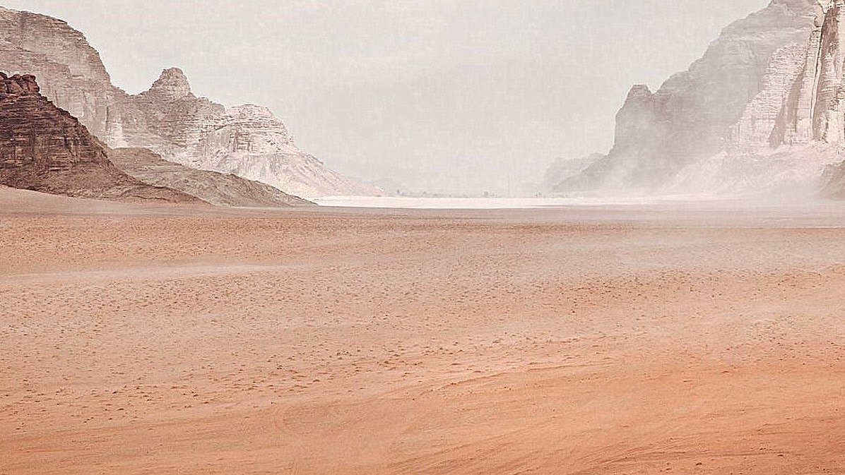 L'image montre un vaste paysage désertique. On peut voir des montagnes rocheuses au loin, entourées de dunes de sable. Le ciel est nuageux, ce qui crée une atmosphère légèrement brumeuse. Au centre, une seule silhouette humaine, probablement un cavalier, se déplace à travers ce paysage aride. La scène transmet une sensation d'immensité et de solitude.
