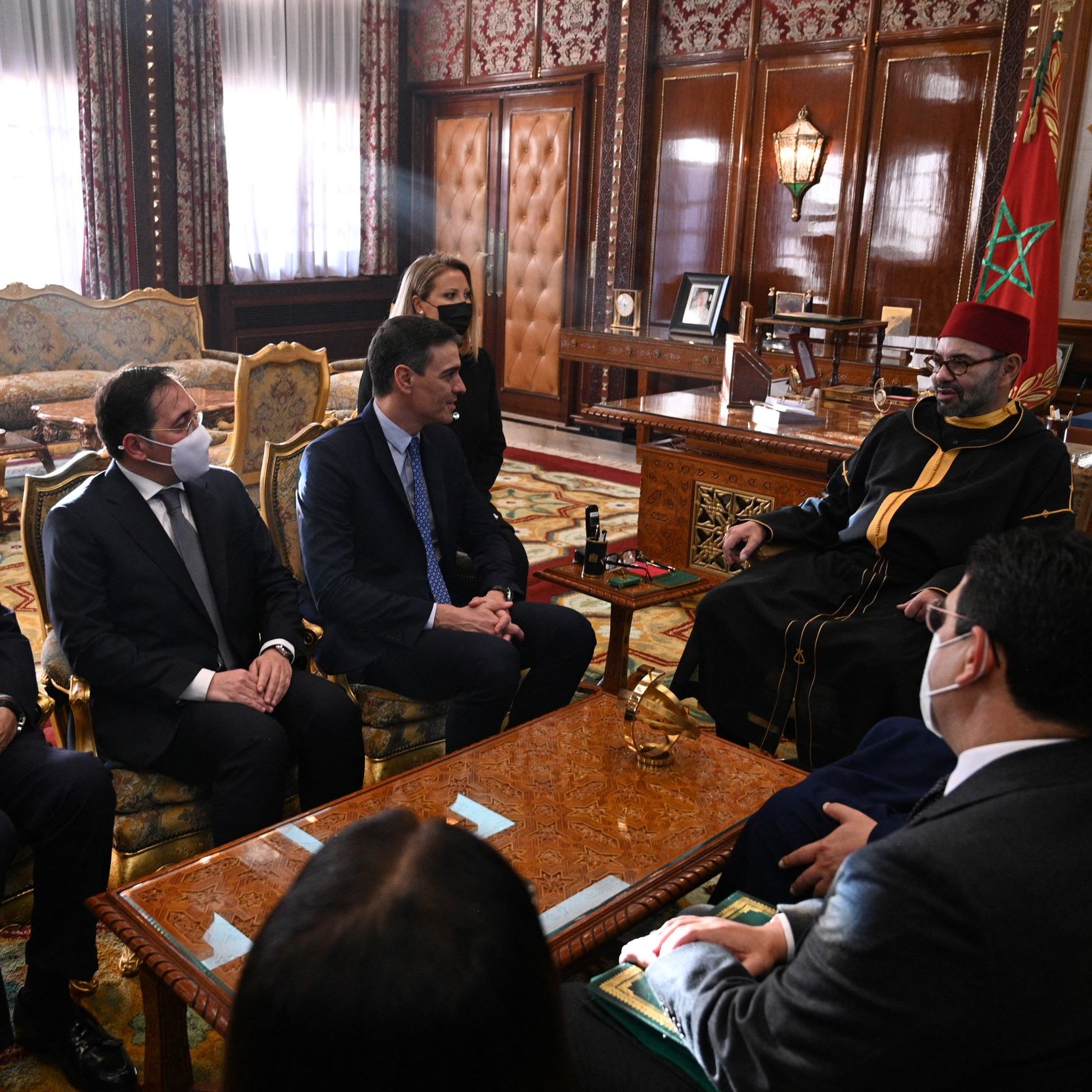 L'image montre une réunion dans un cadre officiel, probablement un bureau ou une salle de réunion importante. Plusieurs personnes en costumes sont assises autour d'une table, tandis qu'une autre personne, vêtue d'une tenue traditionnelle, est assise en face d'eux. Les participants portent des masques, ce qui indique que la réunion pourrait avoir lieu en période de pandémie. L'environnement est richement décoré, avec des éléments de design marocain et un mobilier en bois orné.