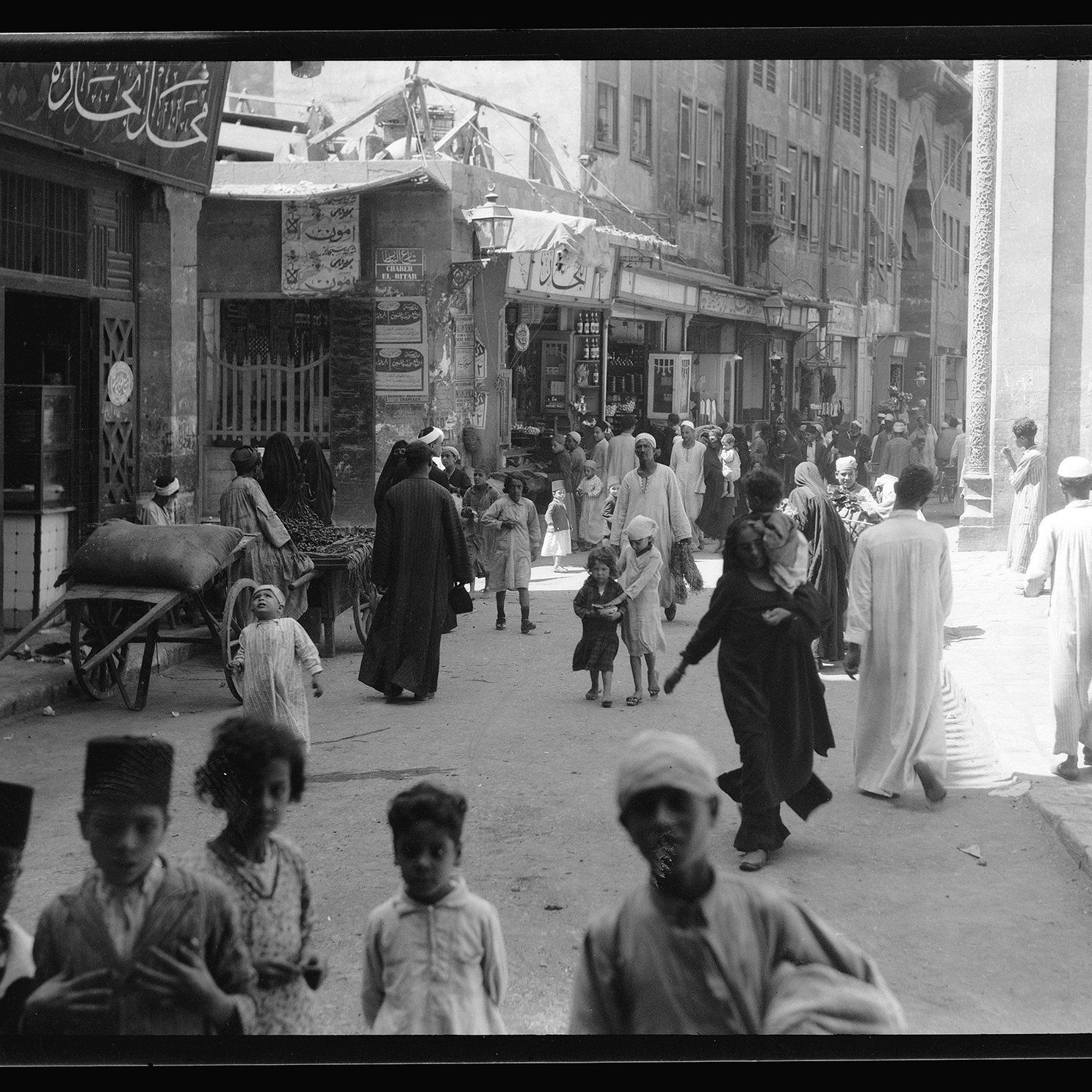 L'image montre une scène animée d'une rue dans une ville. On y voit des groupes de personnes marchant, certaines en conversation. Des enfants jouent au milieu des passants. L'architecture autour d'eux suggère un paysage urbain traditionnel, avec des bâtiments ornés et des boutiques. Il y a aussi une charrette et des femmes et hommes vêtus de vêtements traditionnels. L'ambiance générale semble dynamique et vivante, reflétant la culture locale.