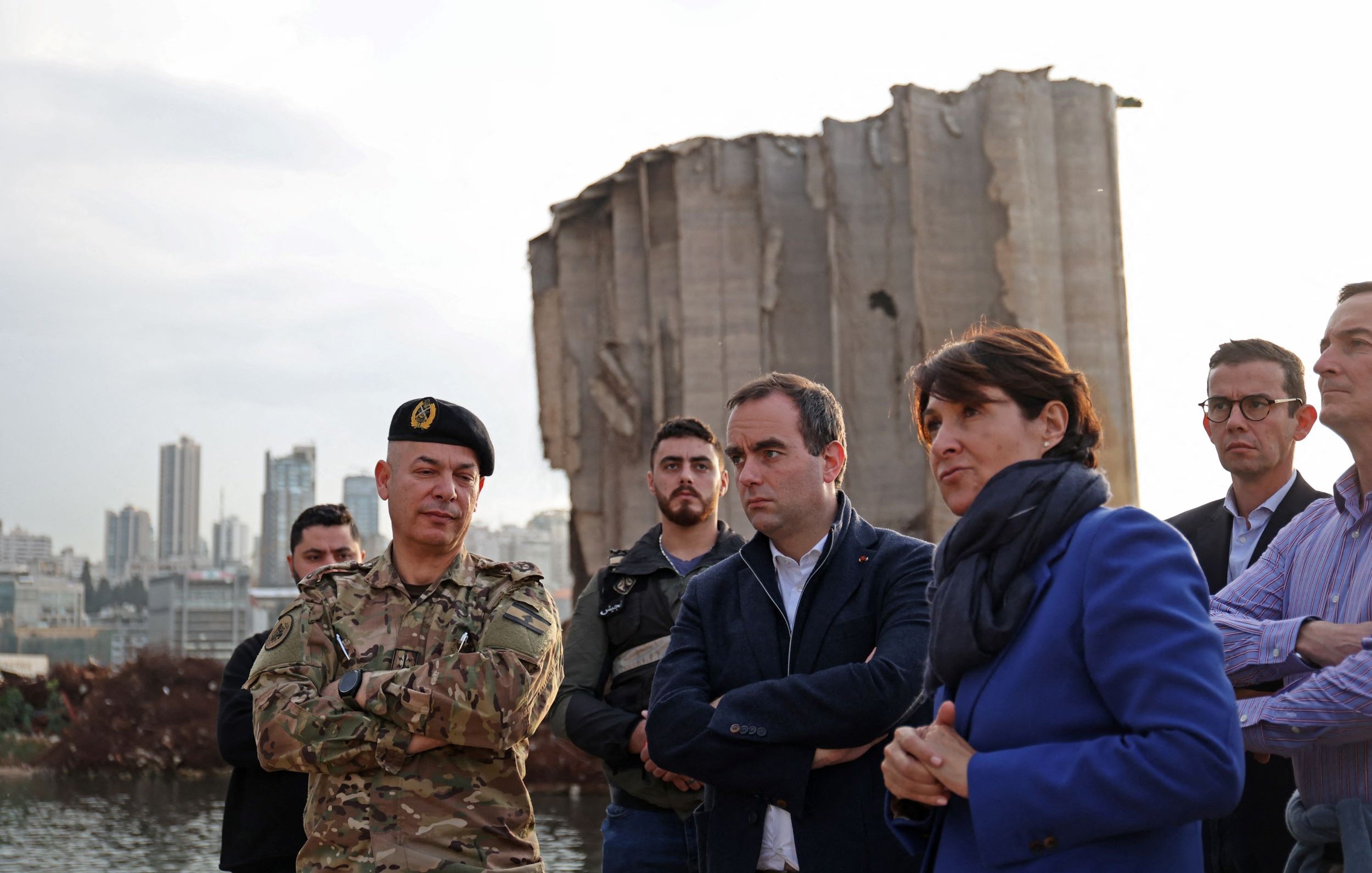 در این تصویر، گروهی از افراد در کنار یک ساختمان ویران، که ظاهراً بخشی از آن تخریب شده، ایستاده‌اند. یک نظامی با لباس نظامی و کلاه فرماندهی در سمت چپ قرار دارد. کنار او مردان و زنانی با لباس‌های رسمی و غیررسمی ایستاده‌اند و به نظر می‌رسد که در حال صحبت و تبادل نظر هستند. پس‌زمینه تصویر نشان‌دهنده شهری با ساختمان‌های بلند است. نور در تصویر کم است و فضای کلی حالت جدی و تاملی دارد.