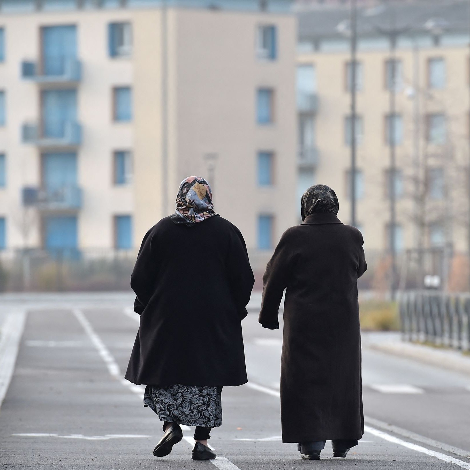 L'image montre deux femmes marchant côte à côte sur une voie urbaine. Elles portent des manteaux sombres et des foulards, ce qui suggère qu'il fait frais. En arrière-plan, on distingue des bâtiments modernes avec des balcons et des fenêtres. La scène dégage une atmosphère tranquille, avec un chemin bordé d'herbes. Leurs silhouettes semblent illustrer un moment de complicité et de camaraderie.