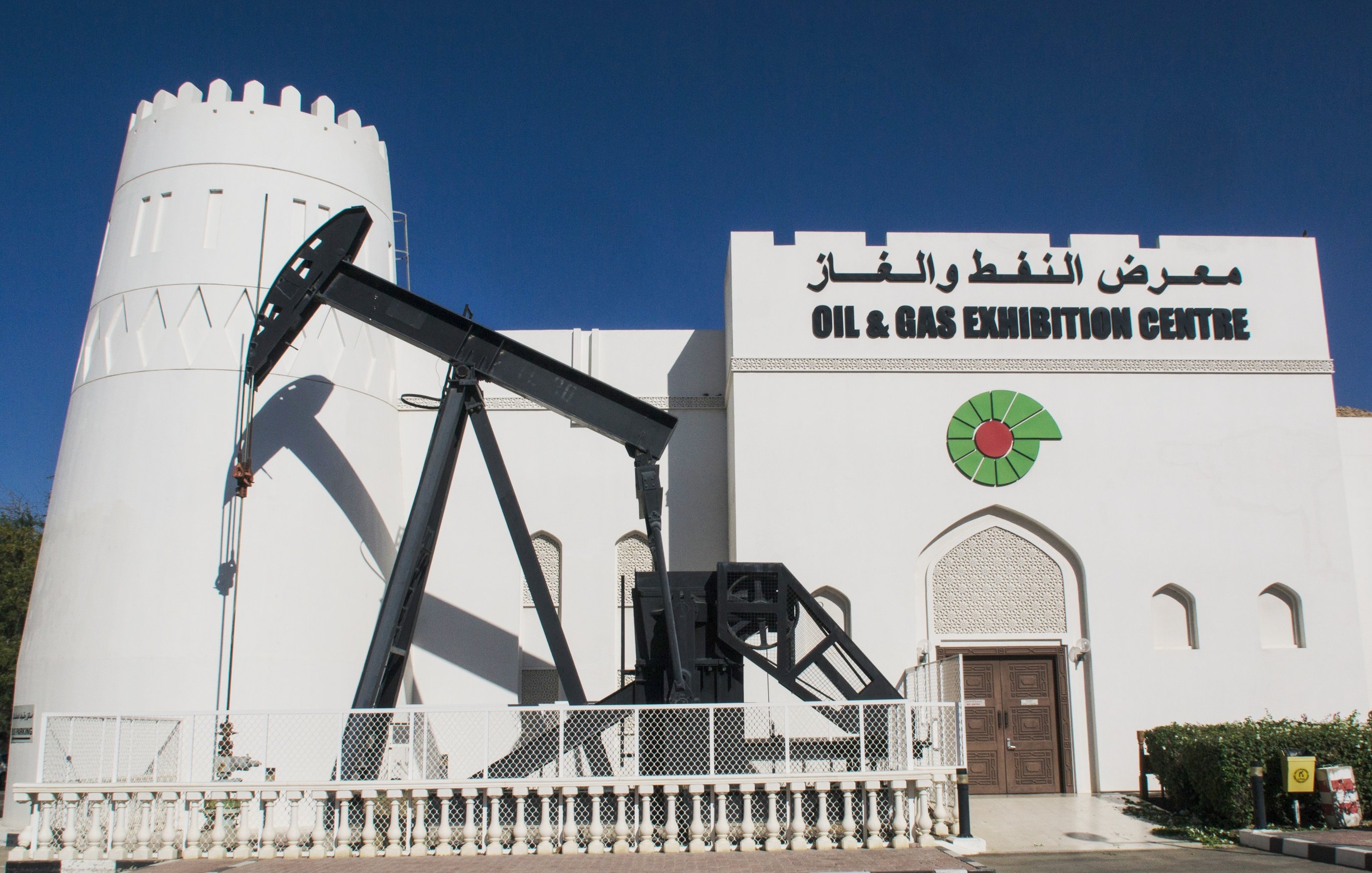 الصورة تظهر مبنى يبدو كمعرض للنفط والغاز، حيث يتضمن تصميمه معالم معمارية مميزة مثل جدران بيضاء وأبراج. أمام المبنى يوجد جهاز استخراج النفط المعروف بـ "رافعة النفط" الذي يُستخدم في عمليات الحفر والتنقيب عن النفط. كما يُلاحظ في التصميم وجود شعار معبر يحتوي على رمز يشير إلى الطاقة، مع كلمات تشير إلى أهمية المعرض في مجال النفط والغاز. السماء في الصورة واضحة والجو يبدو مشمسًا.